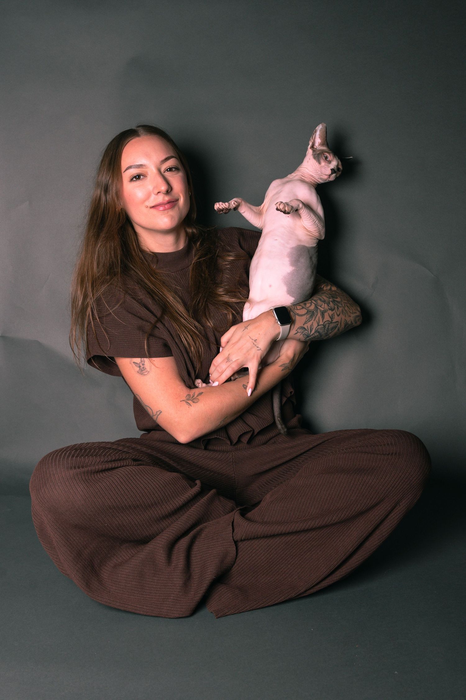 Young woman with tattoos sits cross-legged holding a Sphynx cat, wearing brown outfit against gray background.