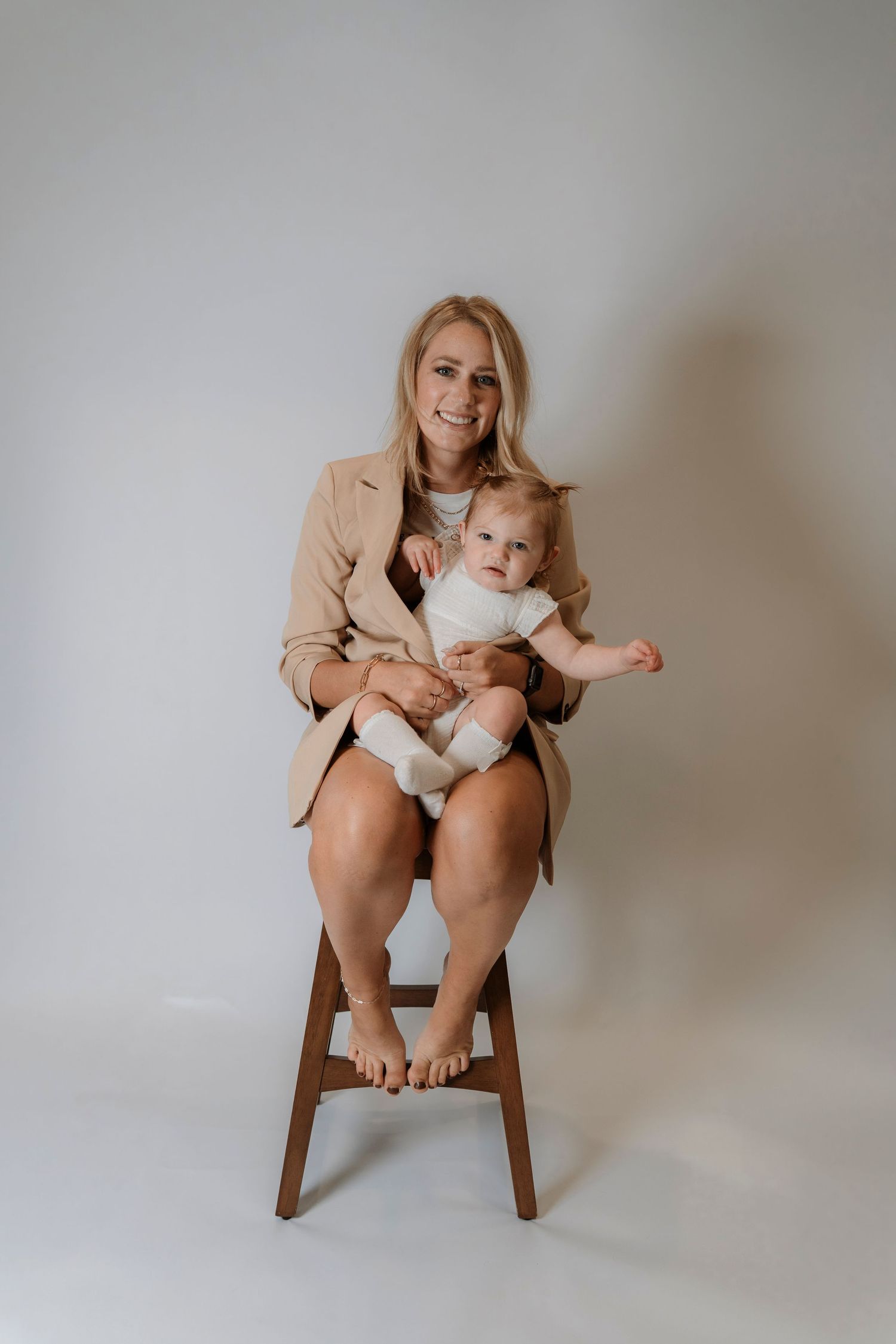 Smiling blonde woman in beige blazer sitting on wooden stool holding baby in white onesie against light background.