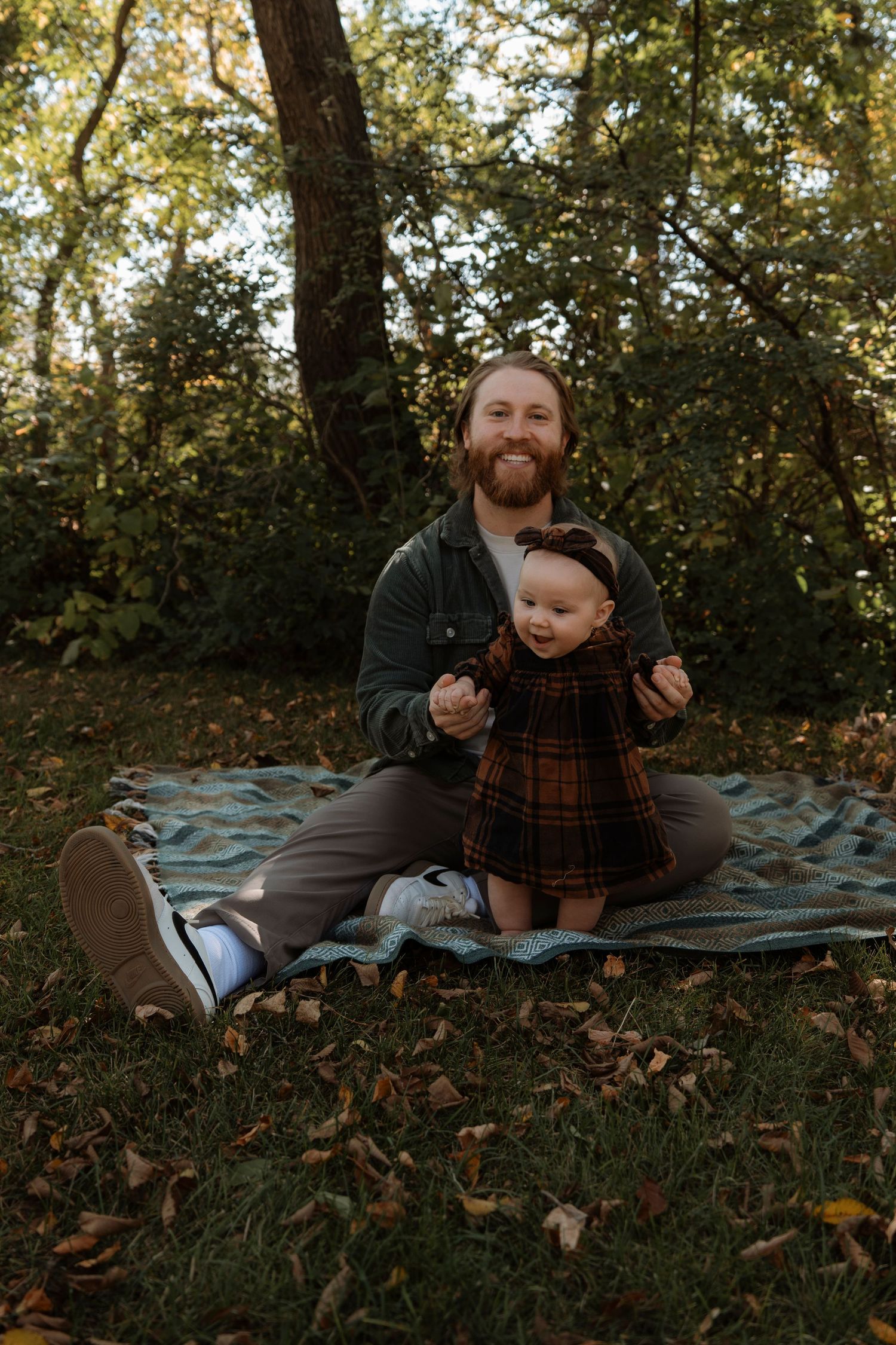 Smiling bearded father sitting cross-legged on blanket outdoors, holding baby girl in plaid dress among autumn leaves.