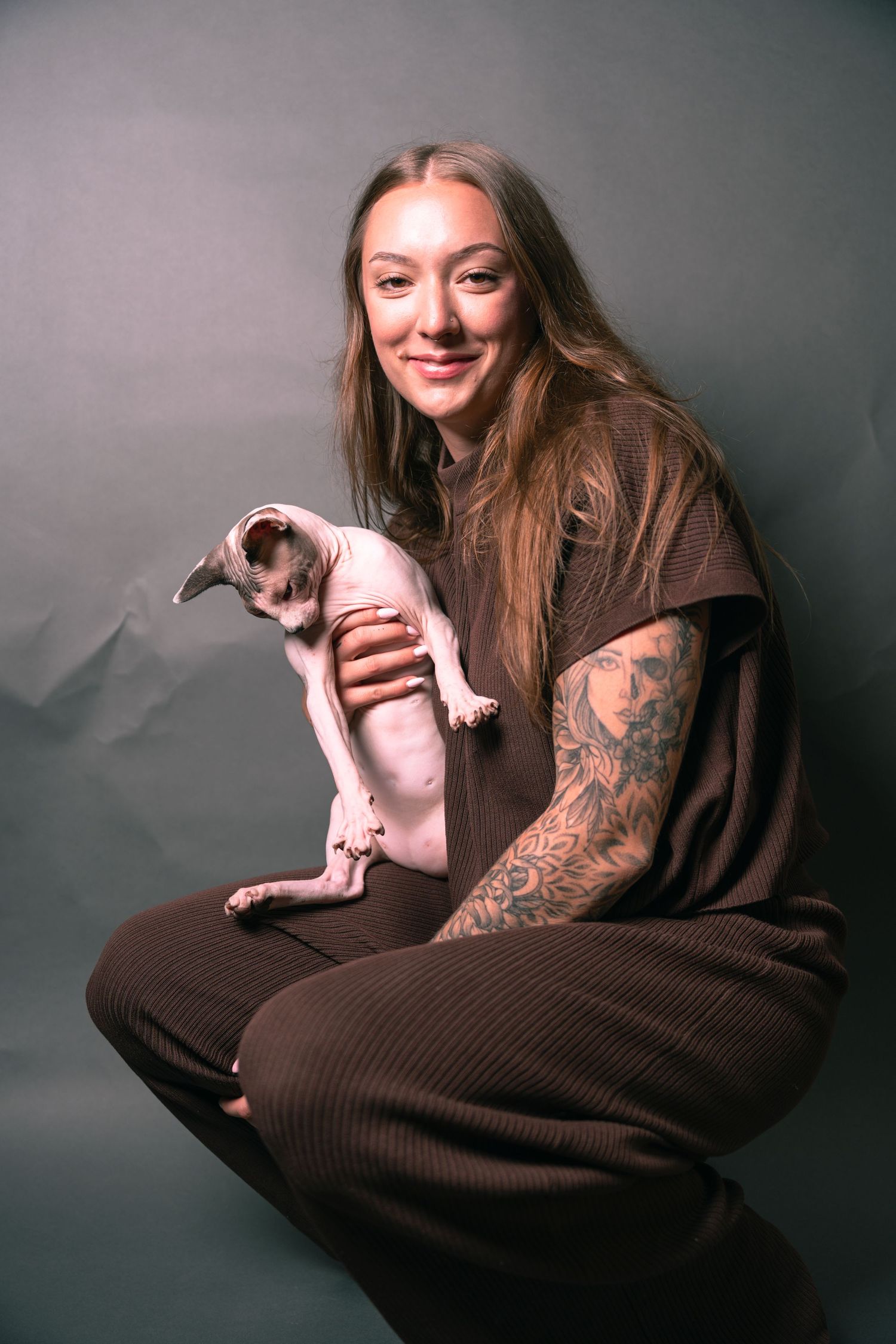 Smiling woman with tattoos crouching and holding a hairless Sphynx cat in a studio setting.