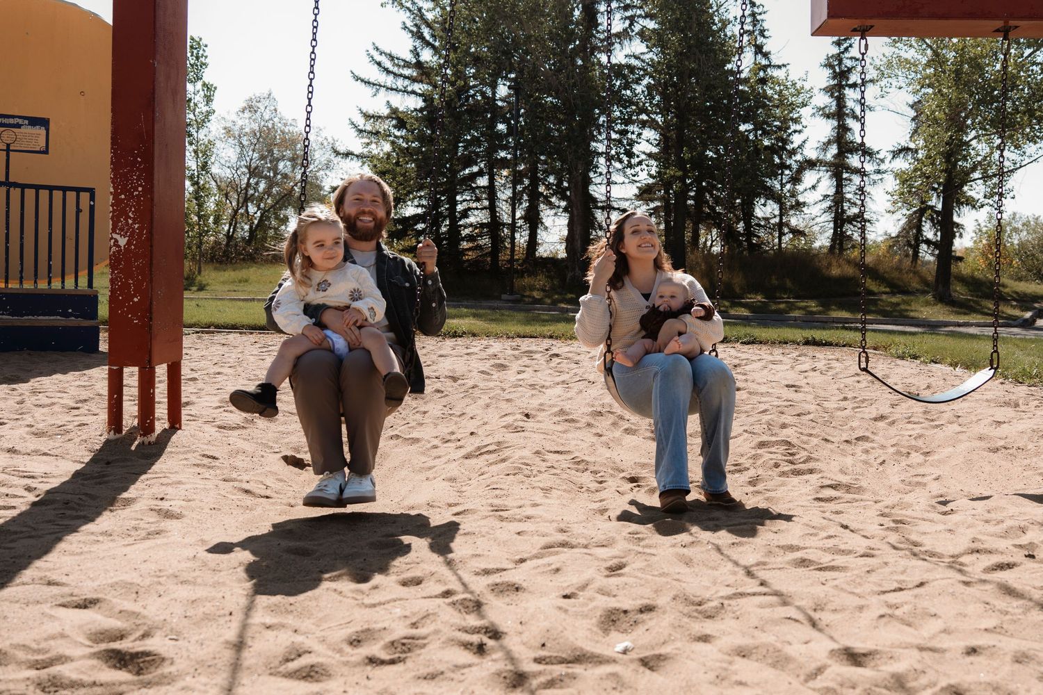 Family with two young children playing on swings at a sandy playground surrounded by tall trees on a sunny day.