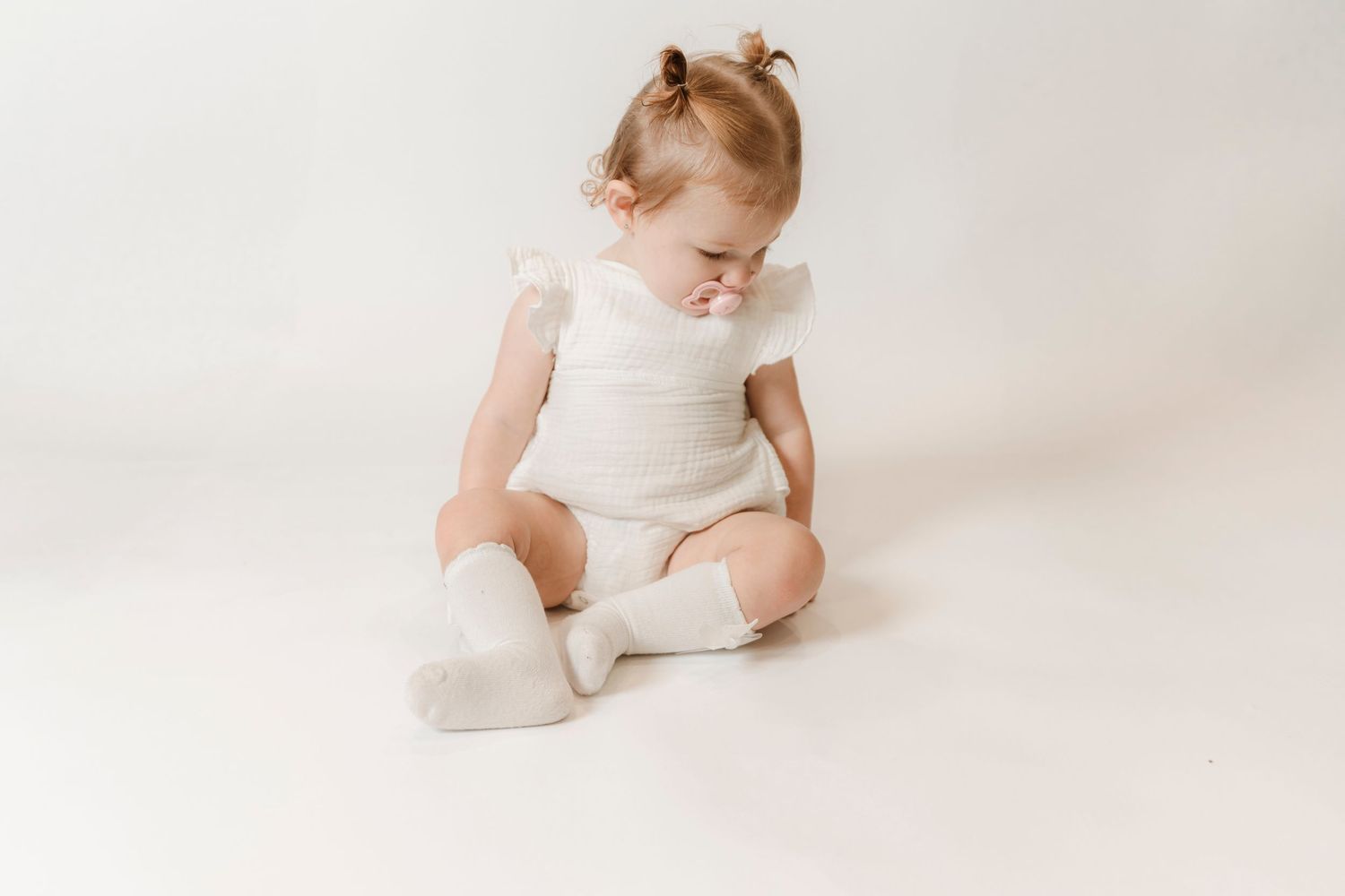 A baby girl in a white romper and knee-high socks sits cross-legged on a white background, looking down.
