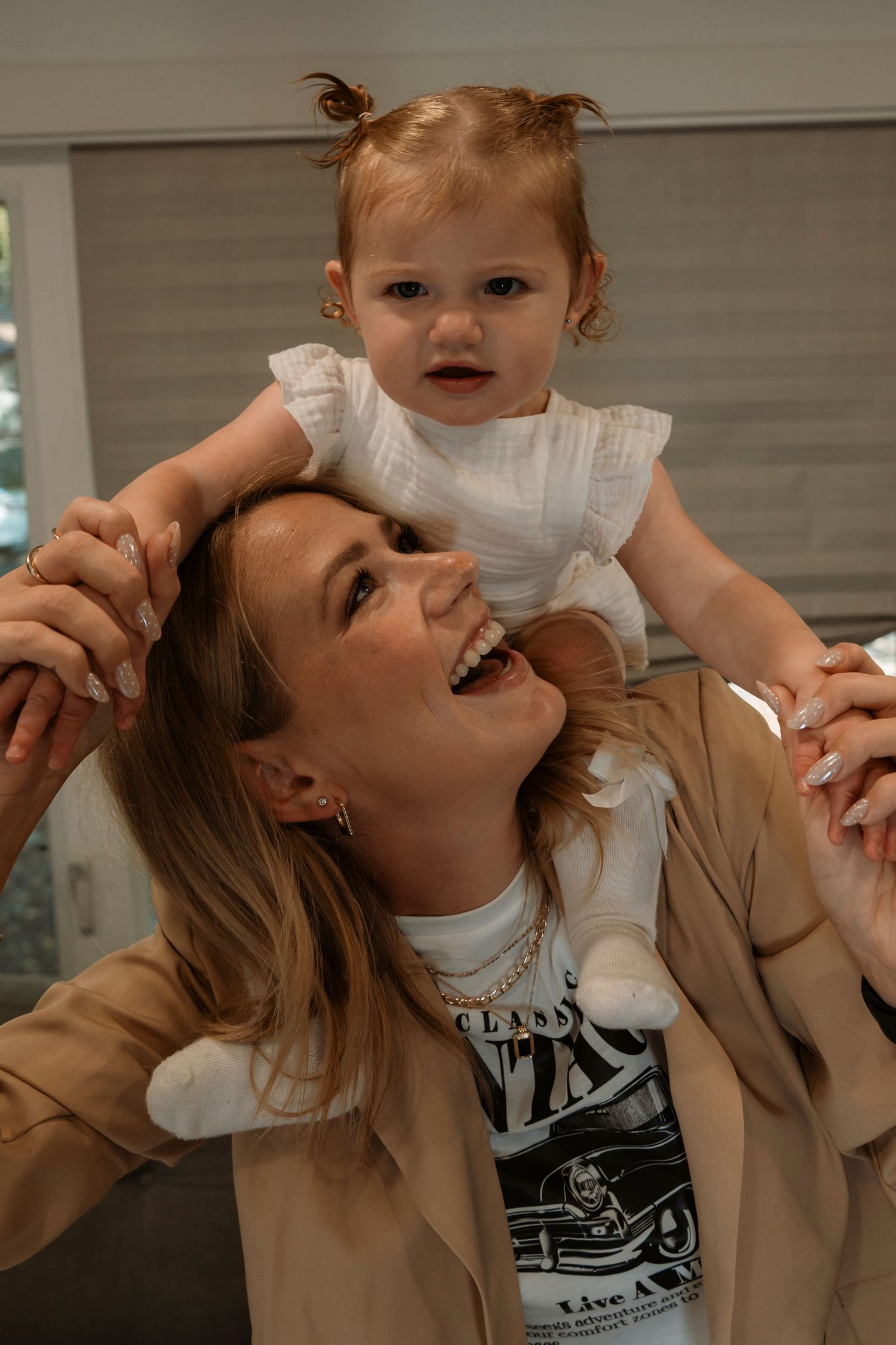 A laughing woman holds a cute baby girl in a white dress on her shoulders indoors.