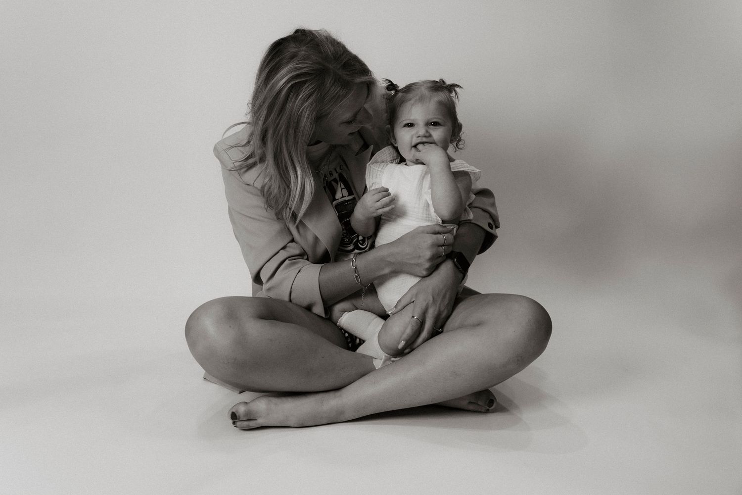 Black and white photo of a mother sitting cross-legged, tenderly kissing her baby girl in a studio setting.