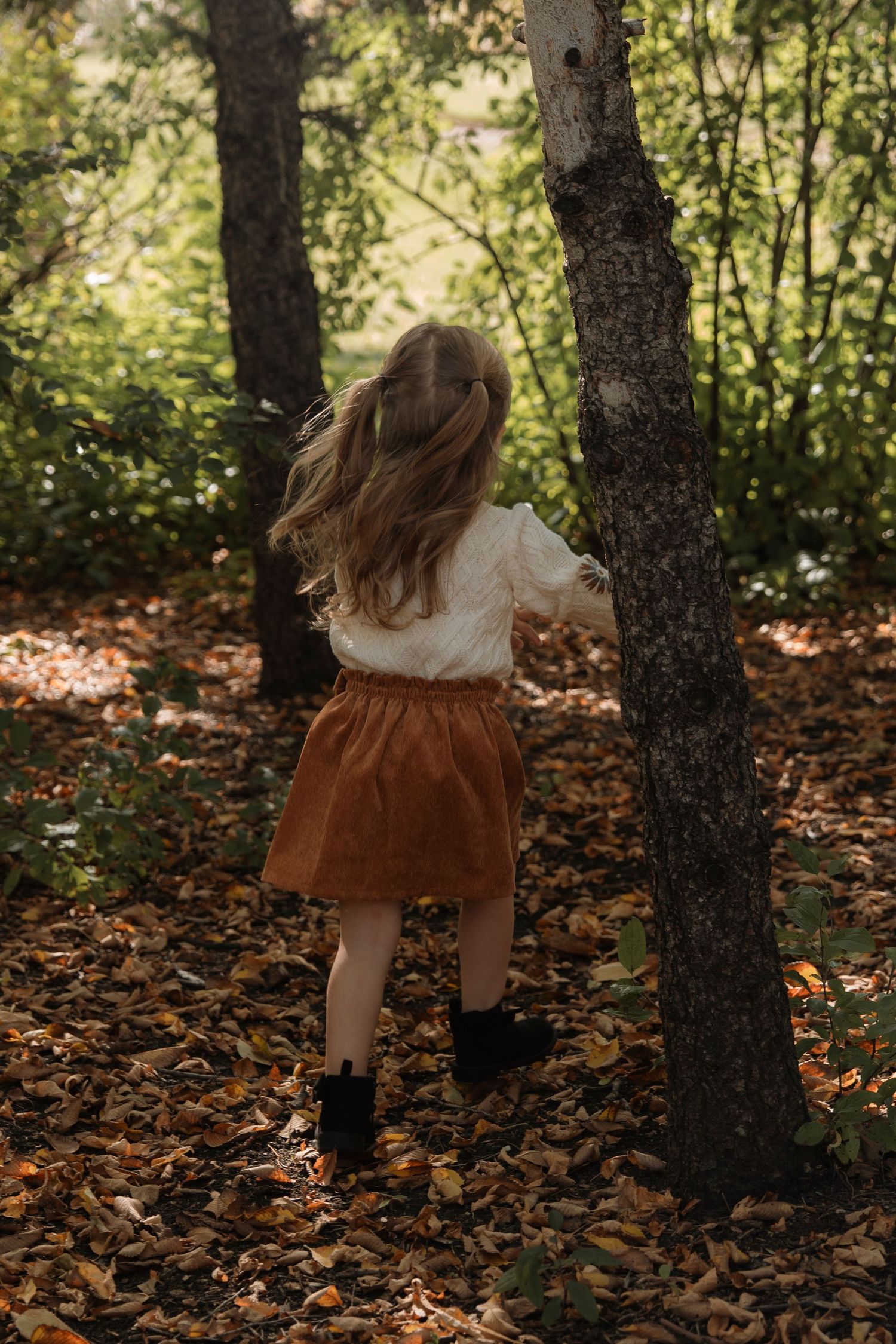 Young girl in white blouse and brown skirt walks through autumn forest with fallen leaves.