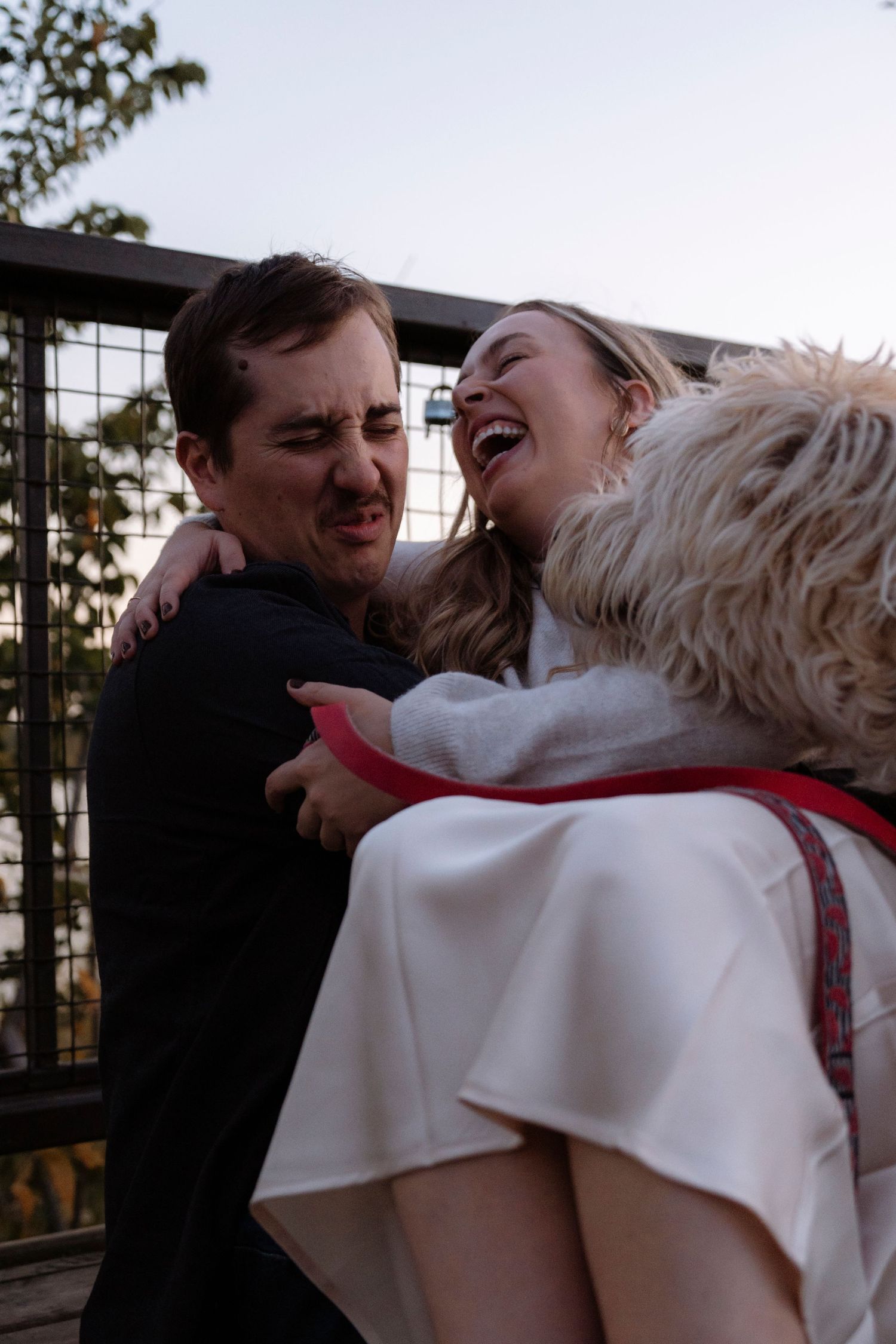A laughing bride in white holds a fluffy dog while embracing a man in black at an outdoor wedding celebration.