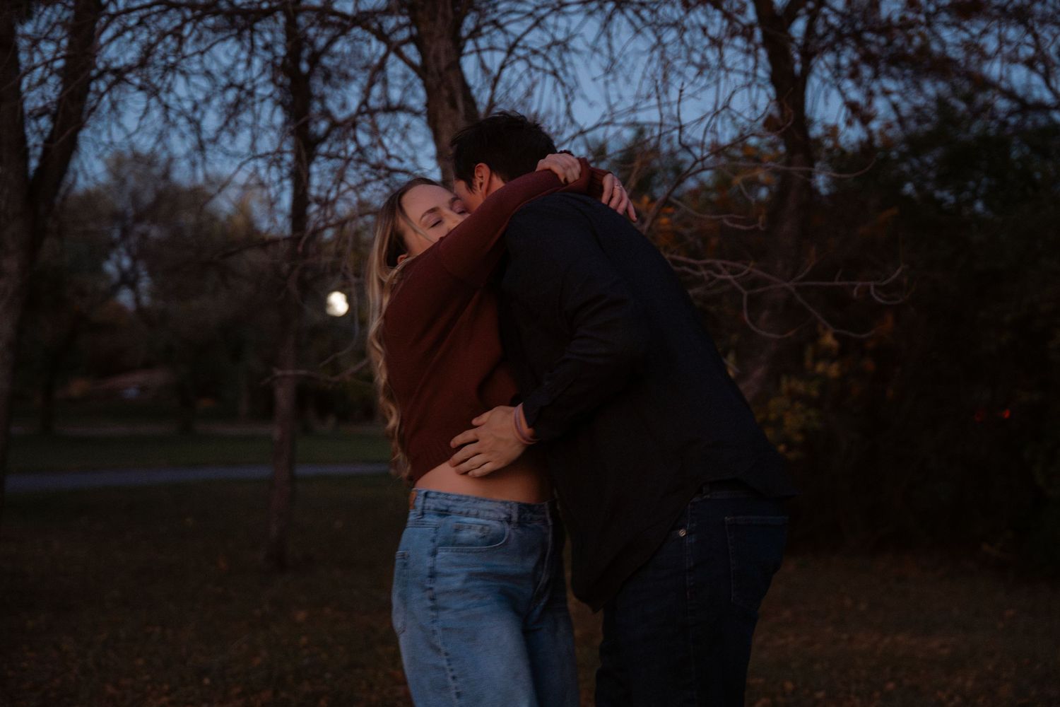 Couple embracing romantically outdoors at dusk with moonrise and bare trees in background.