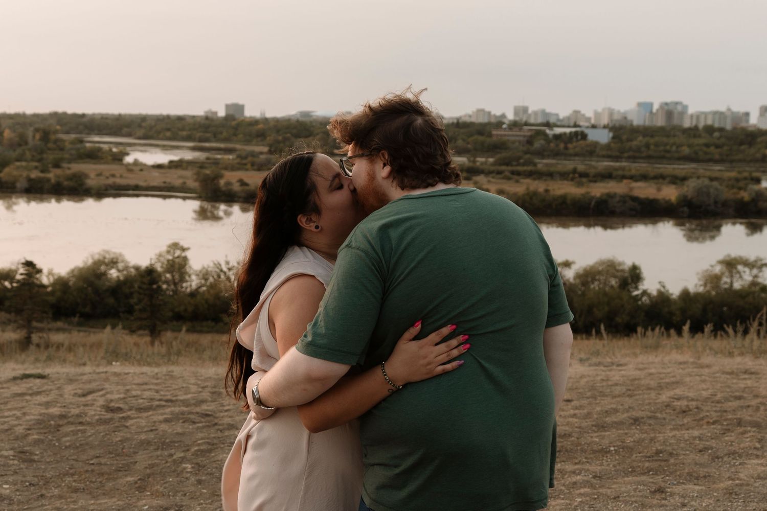 Couple sharing a romantic kiss outdoors near a river at sunset, woman in white dress embracing man in green shirt.