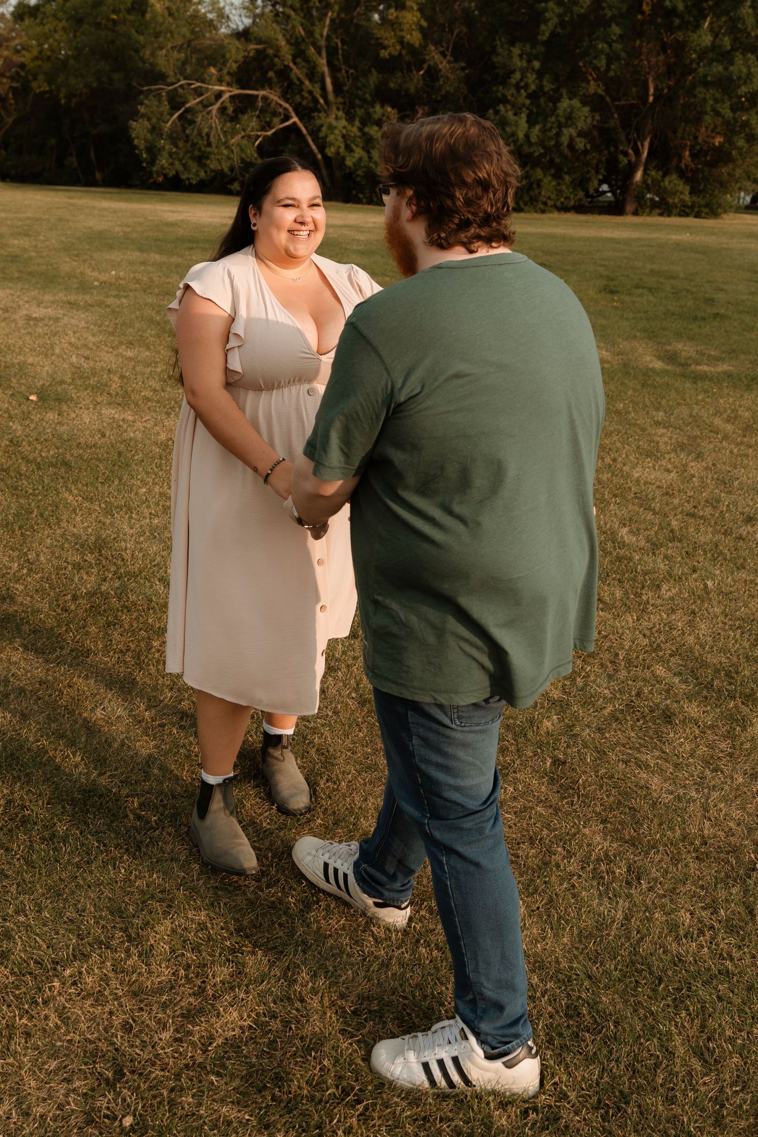 Couple holding hands and smiling in a sunny park, woman in white dress and man in green shirt.