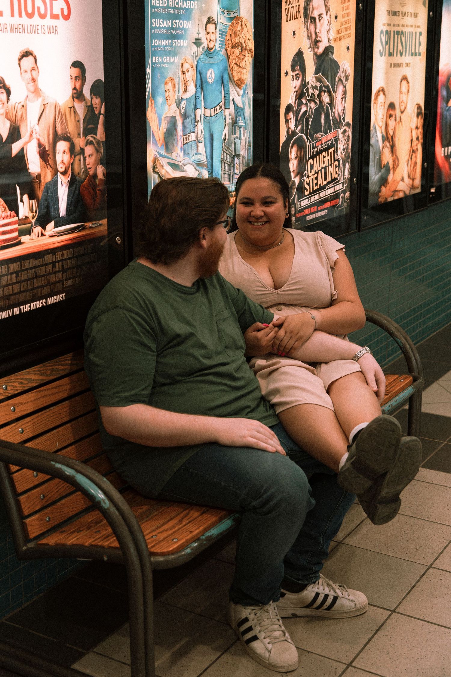 Couple sitting on a bench outside a movie theater, smiling and holding hands in front of film posters.