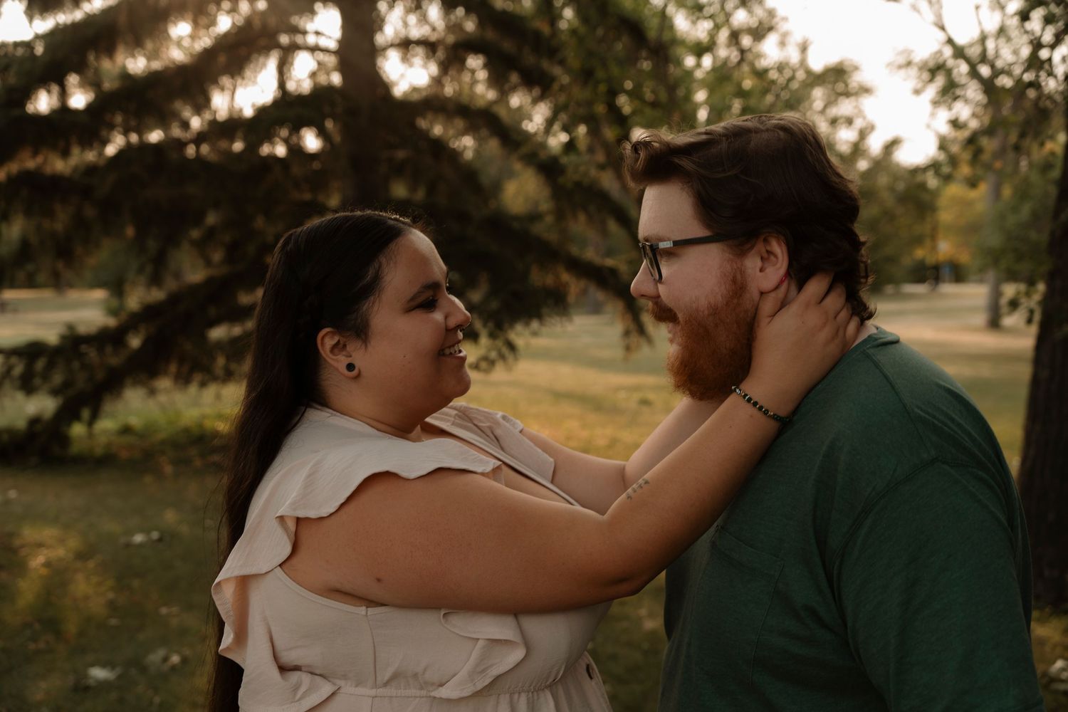 Couple sharing a tender moment outdoors at golden hour, woman in white ruffled top embracing bearded man in green shirt.