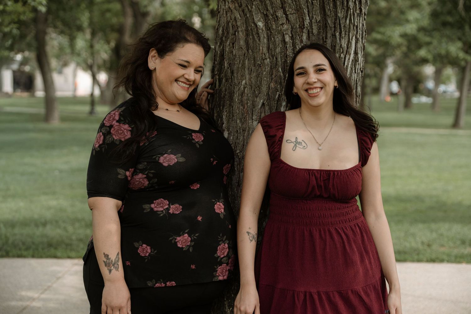 Two women with tattoos smiling outdoors near a tree, one in floral black dress, one in burgundy dress.