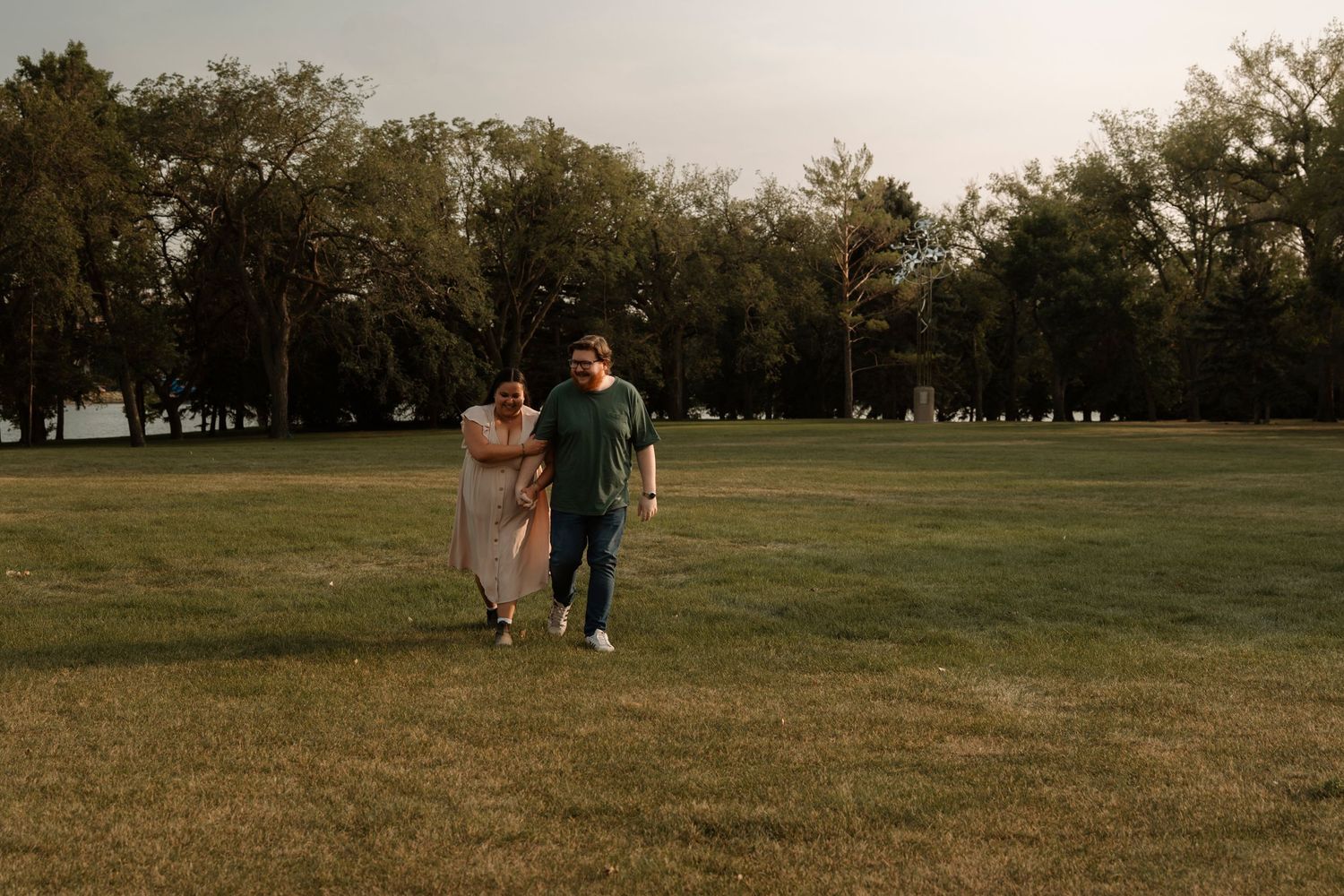 Couple walking hand in hand across a lush green park at golden hour with tall trees in the background.