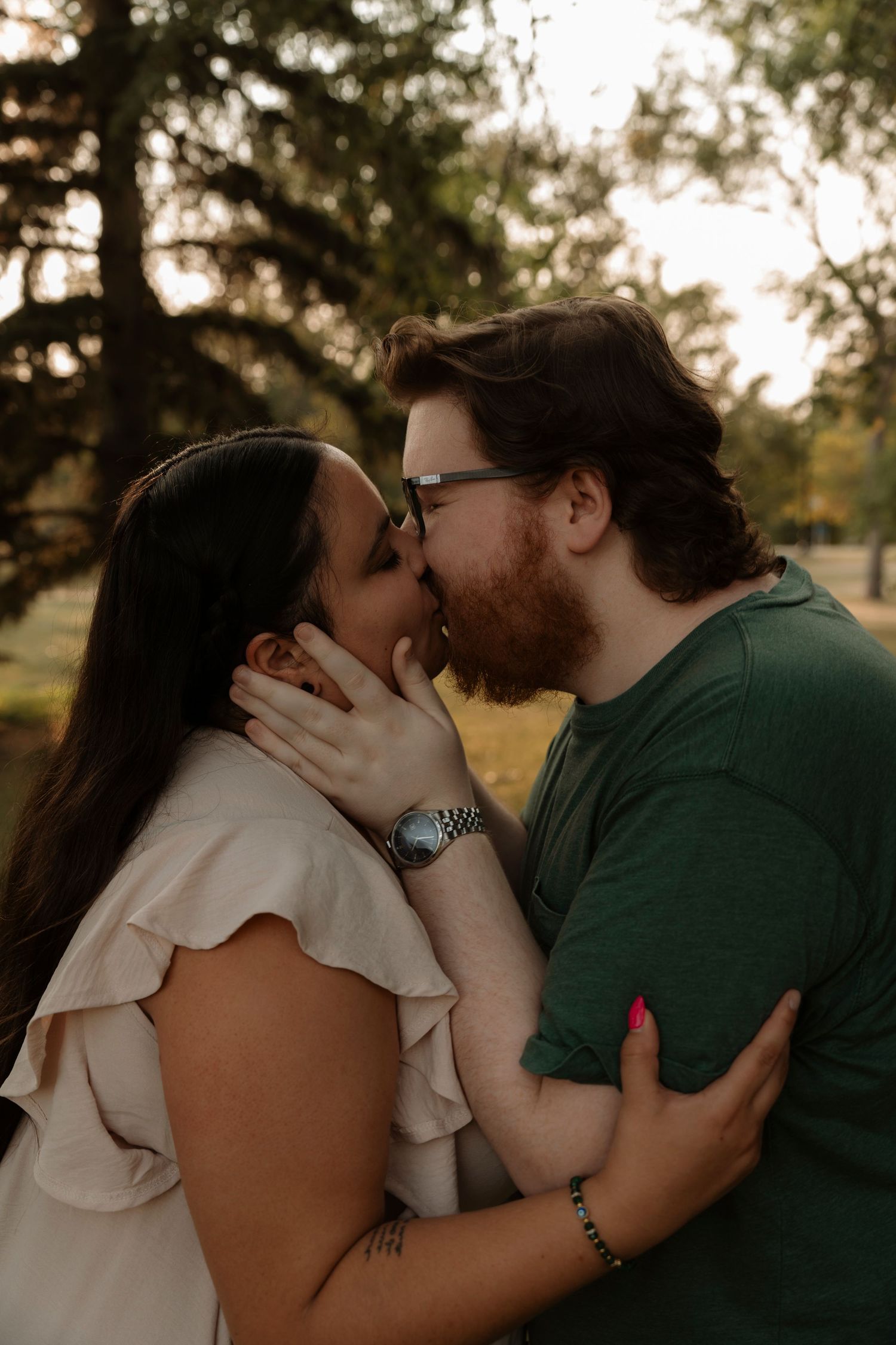 A couple shares a romantic kiss outdoors, man with red beard in green shirt embraces woman in white ruffled top.