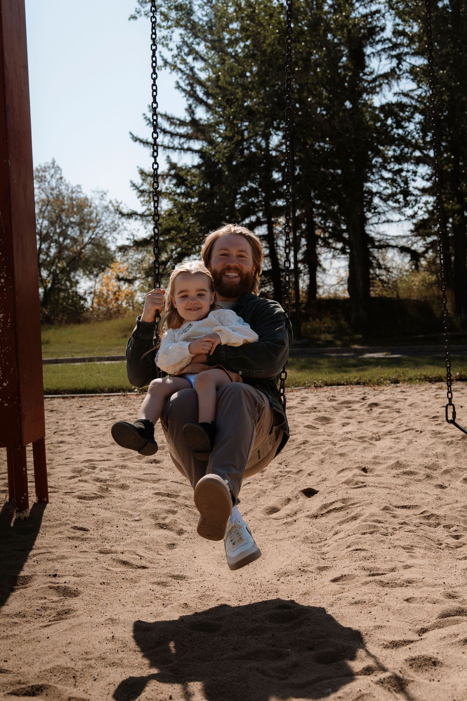 Smiling bearded father holding toddler on playground swing in sandy park surrounded by tall green trees on sunny day.