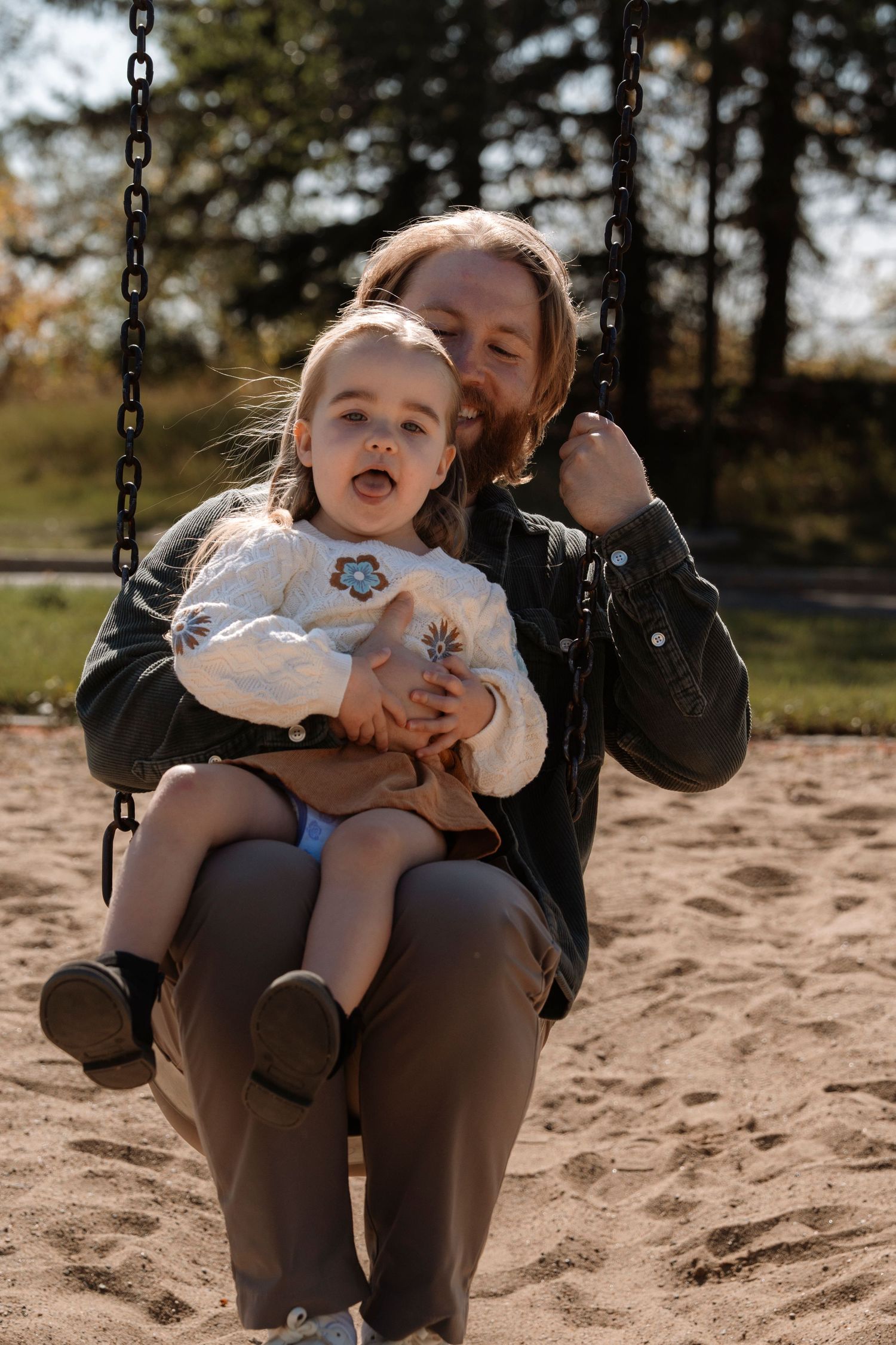 Parent and toddler enjoying a swing together at a sunny playground, surrounded by trees in autumn.