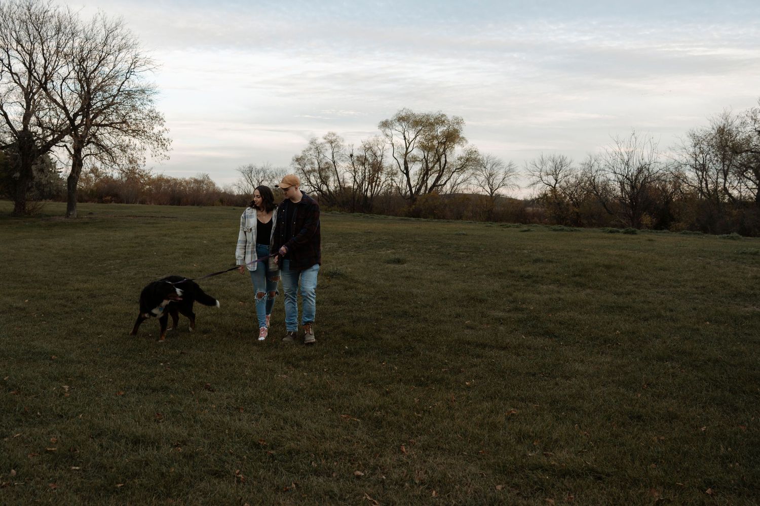 Couple walking a black dog in an open green field with bare trees and overcast sky in autumn.
