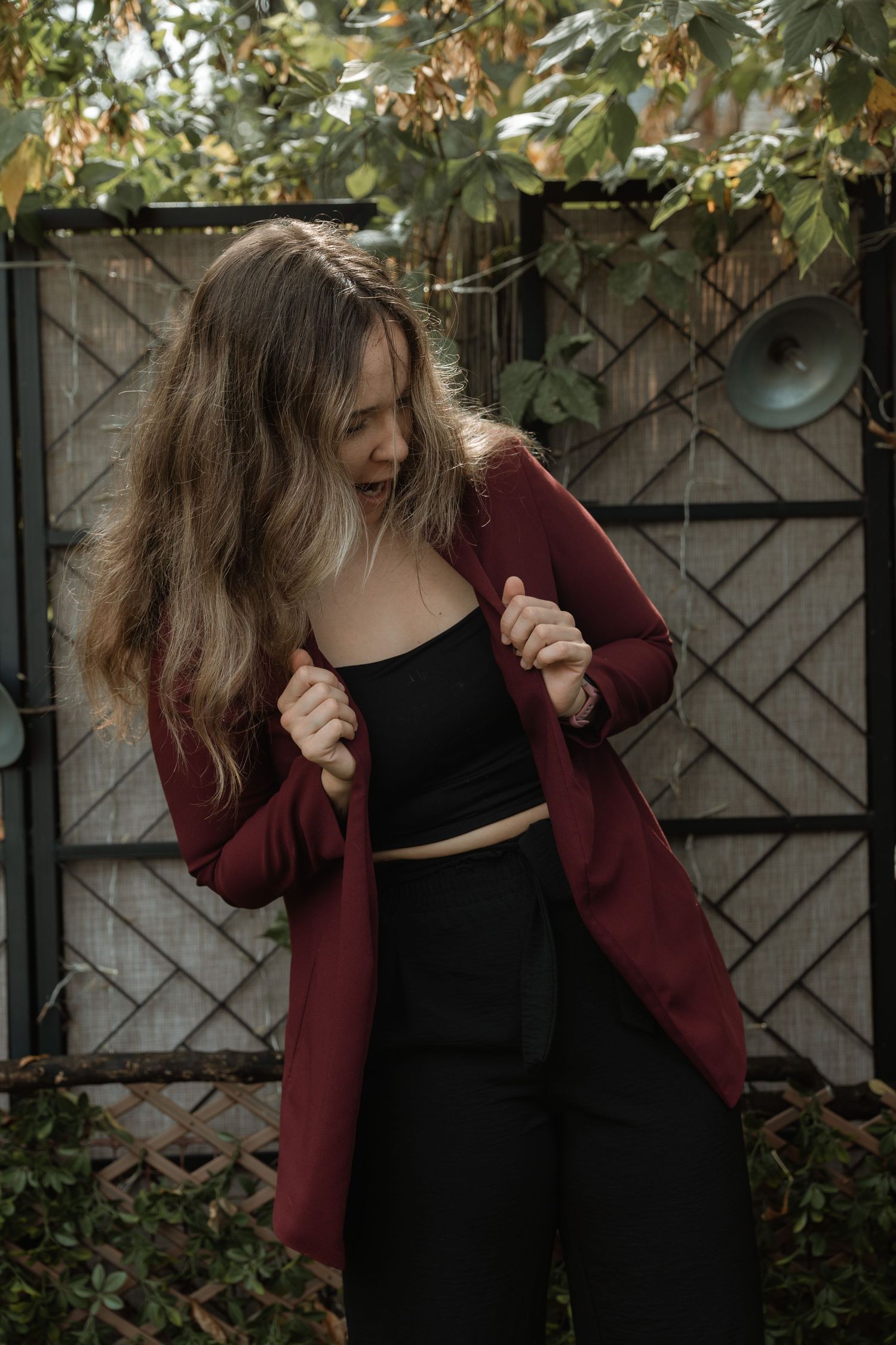A person in a burgundy blazer and black top gestures in an outdoor setting with wooden fence panels.