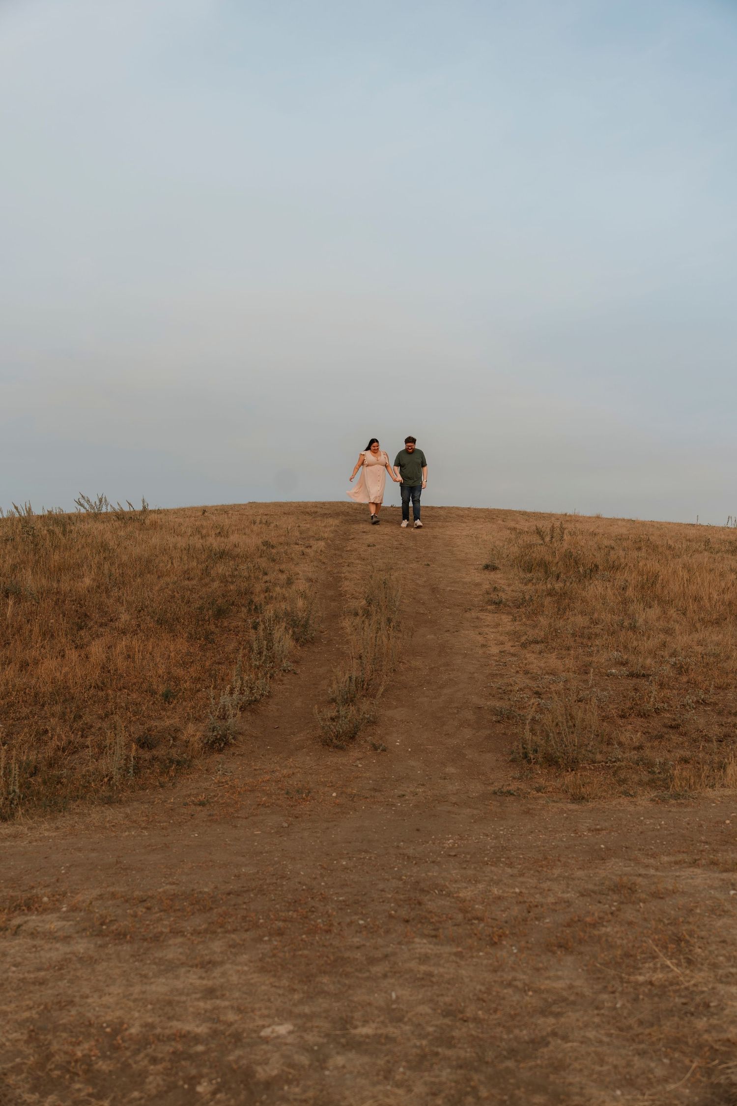 Couple walking hand in hand along a dirt path through golden fields under a hazy sky.