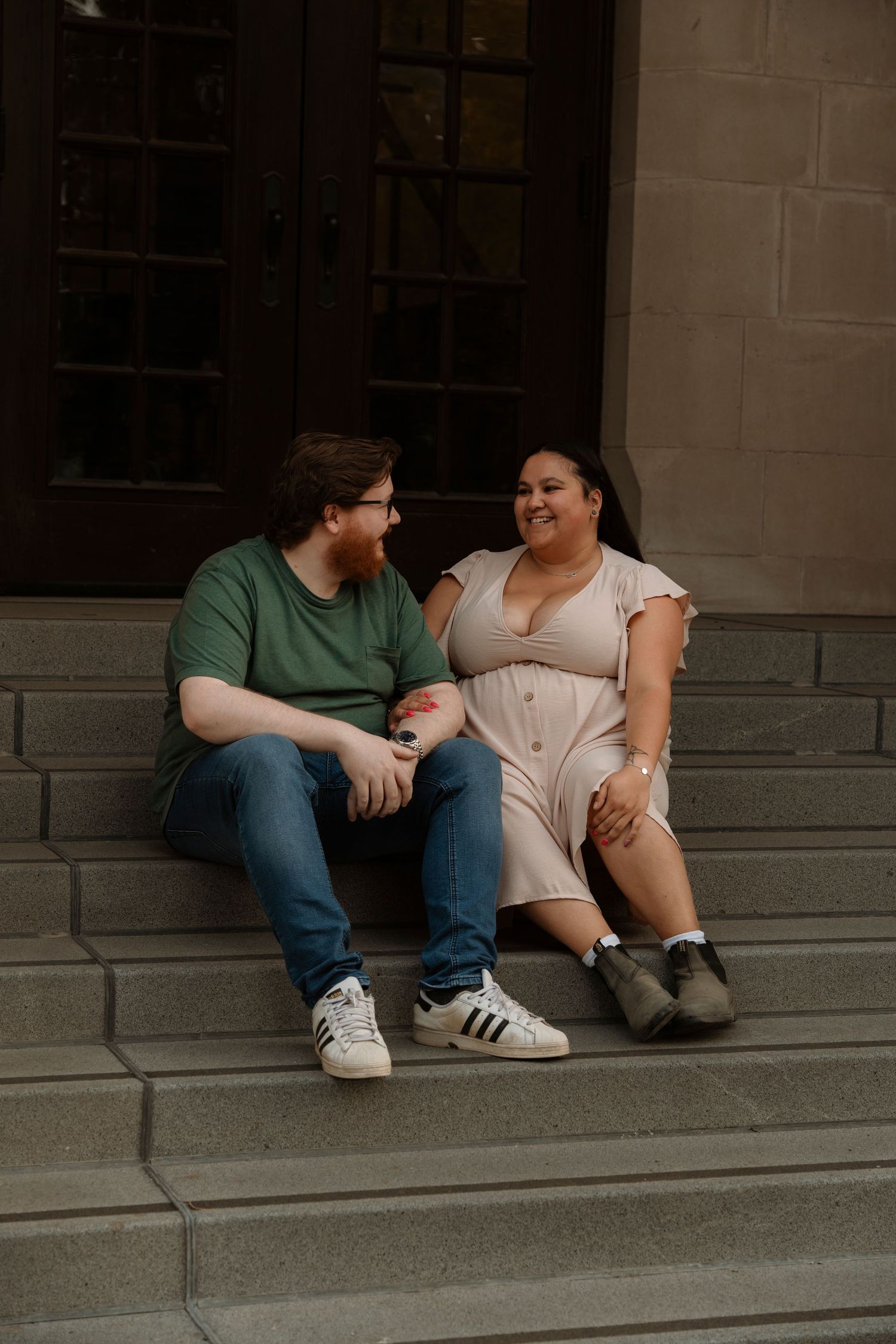 A couple sits laughing together on stone steps, man in green shirt and jeans, woman in pink dress, both wearing sneakers.