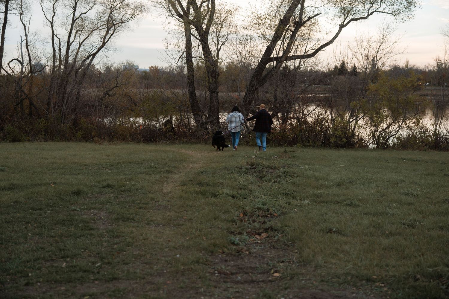 Two people walking a black dog along a grassy riverbank path surrounded by bare trees at dusk.