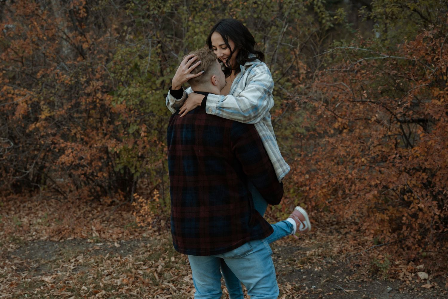 Couple embracing and laughing together in an autumn forest setting, wearing casual flannel and denim outfits.