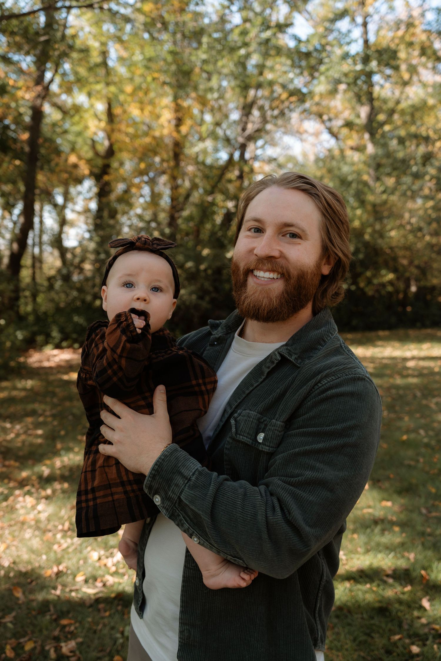 Smiling bearded man holding a baby in a plaid outfit outdoors in a park during autumn.