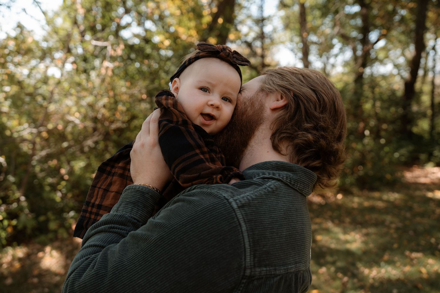 Father kissing baby girl in plaid dress outdoors in autumn forest setting.