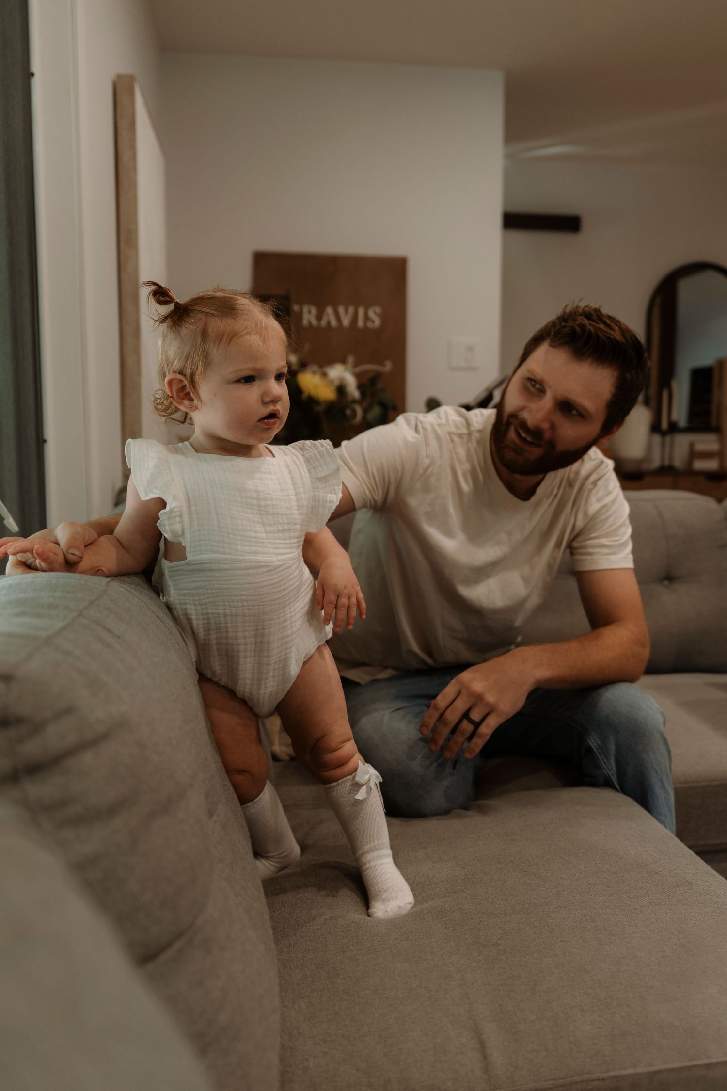 A smiling father watches his toddler daughter stand on a gray sofa in a cozy living room.