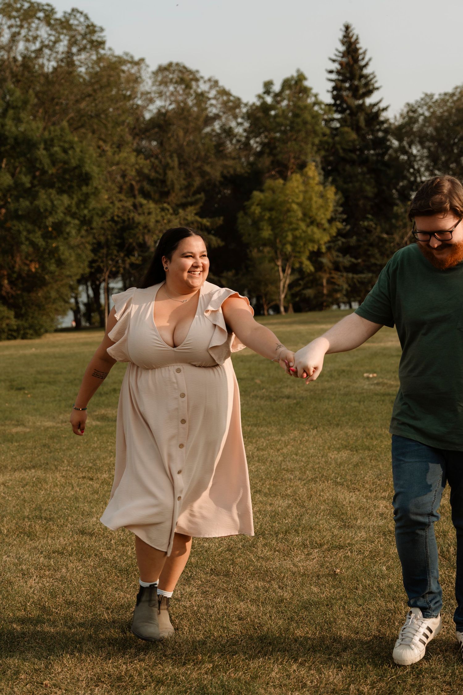 A smiling plus-size woman in a pink dress holds hands with a bearded man in green shirt while walking in a sunny park.