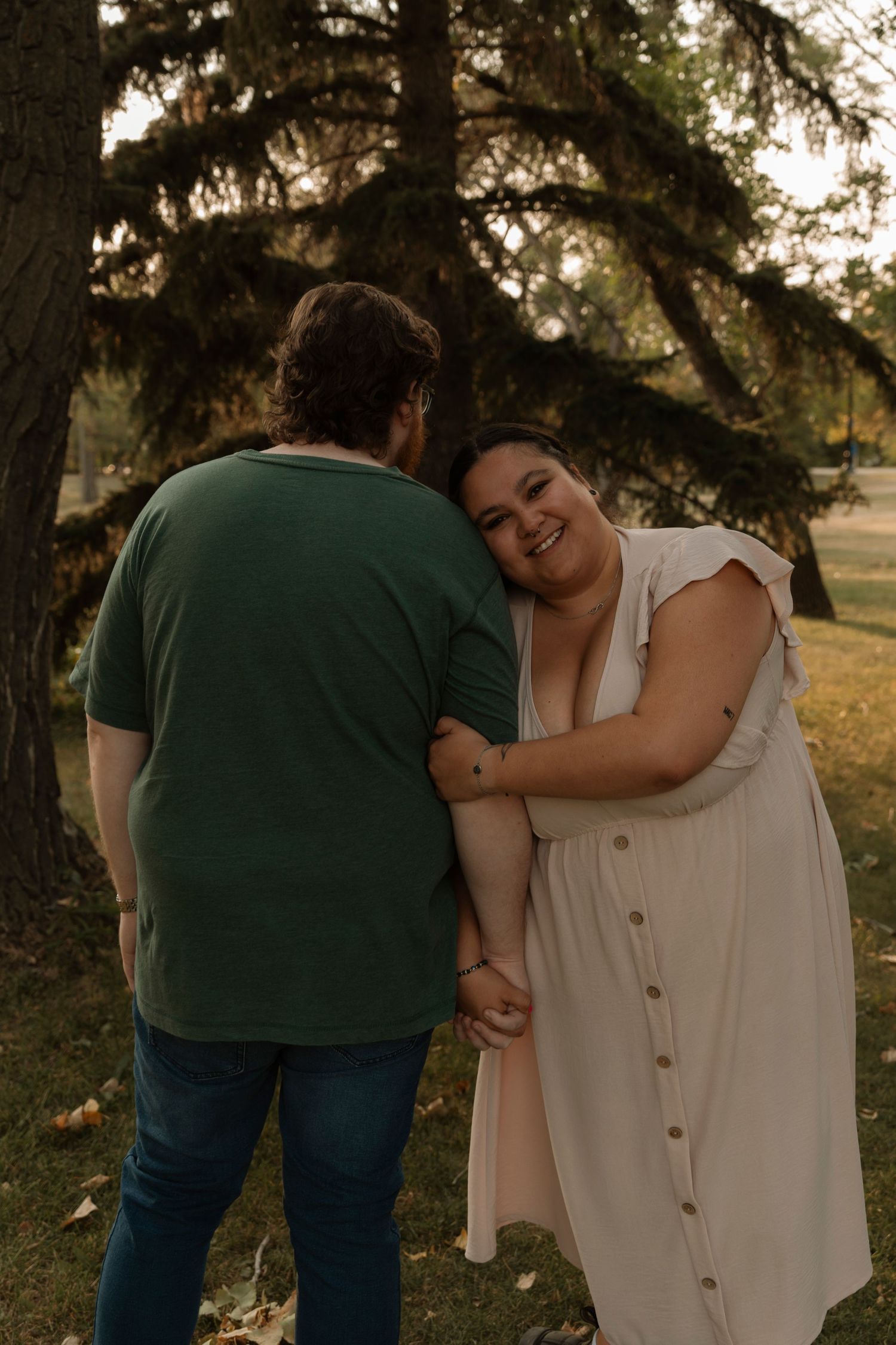 A smiling woman in a cream dress leans on a man in a green shirt outdoors in a park setting.