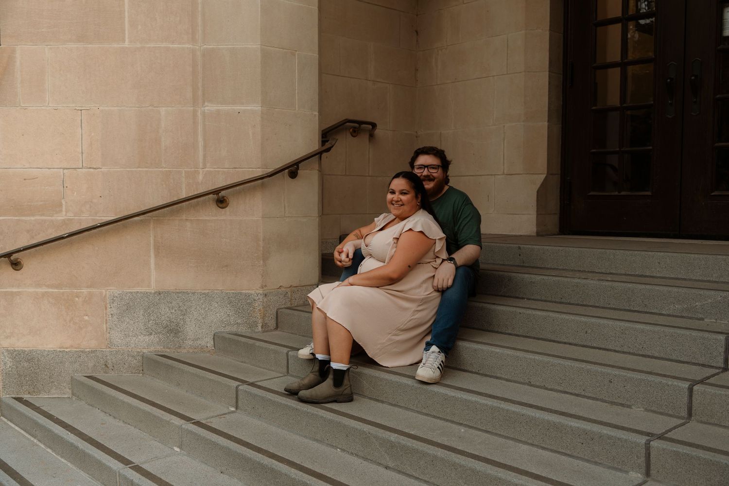 Couple sitting together on stone steps outside a historic building, woman in pink dress and man in green shirt.