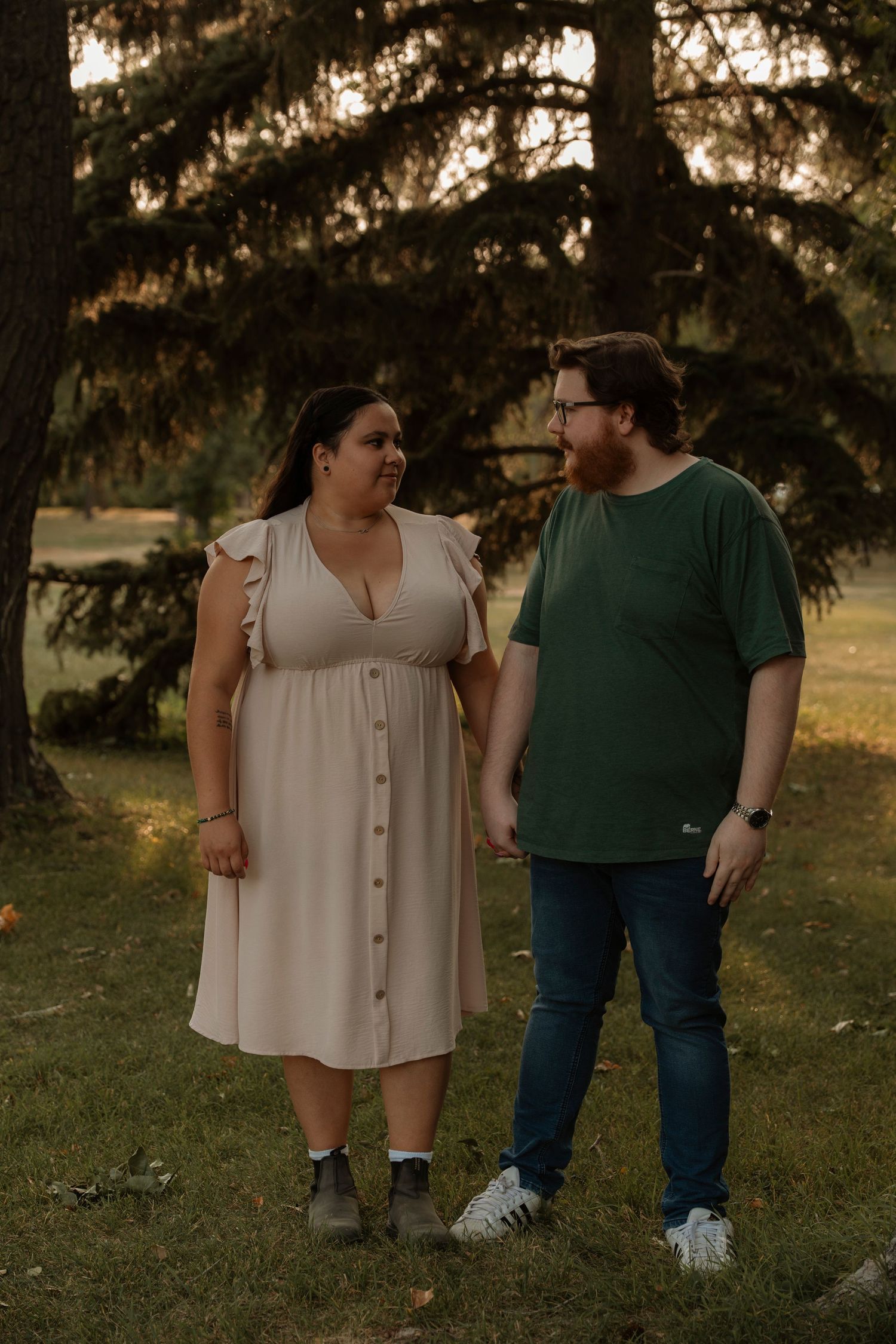 A couple holding hands and smiling at each other in a park, woman in beige dress, man in green shirt and jeans.