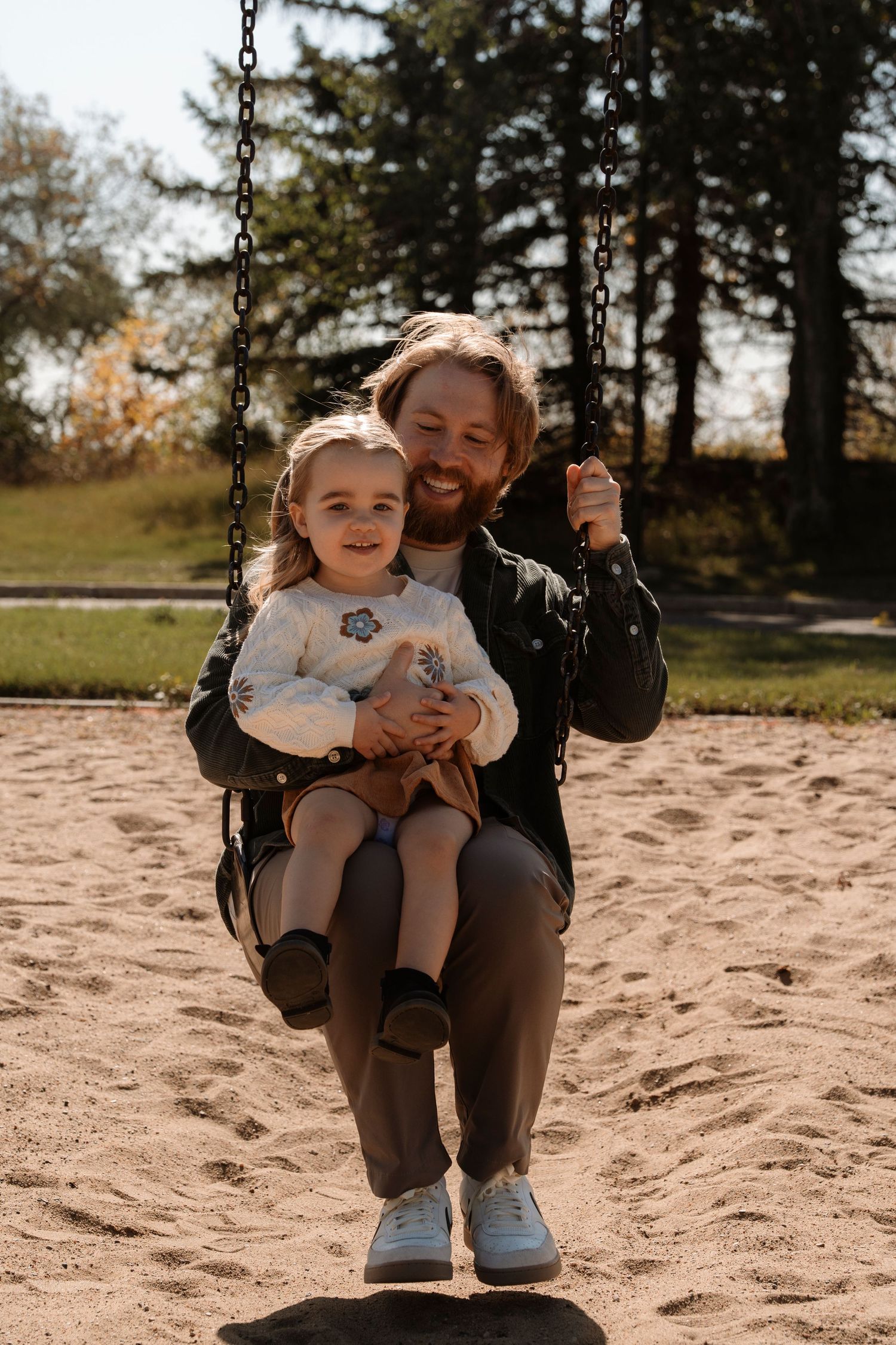A smiling father sits on a playground swing with his young daughter in a sandy park surrounded by autumn trees.
