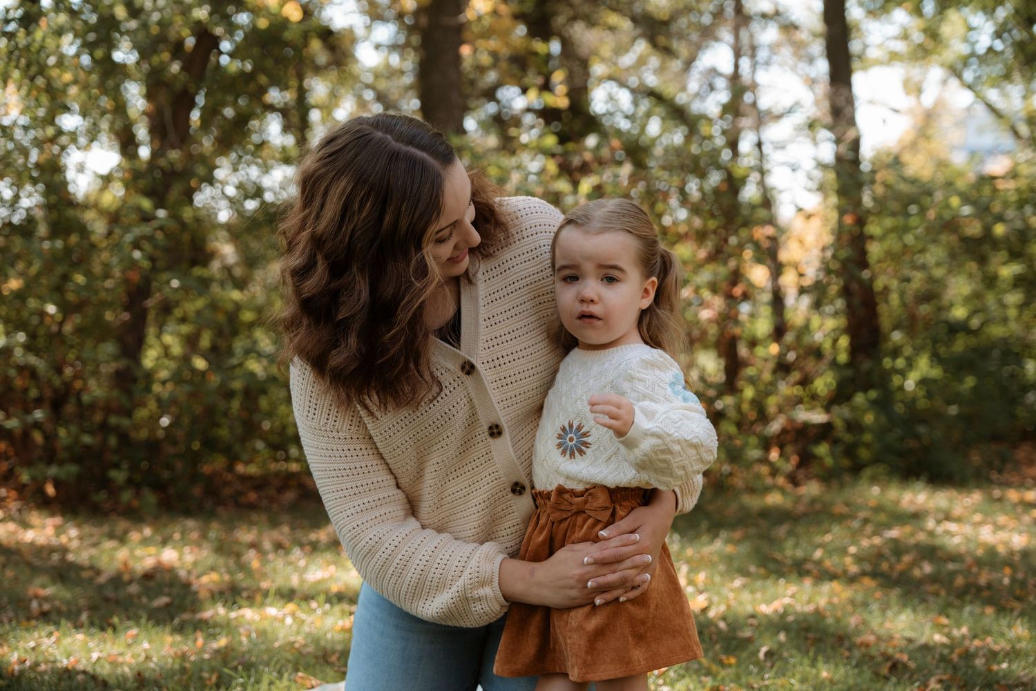 A mother holds her toddler in an autumn forest, both wearing cozy sweaters and warm tones.