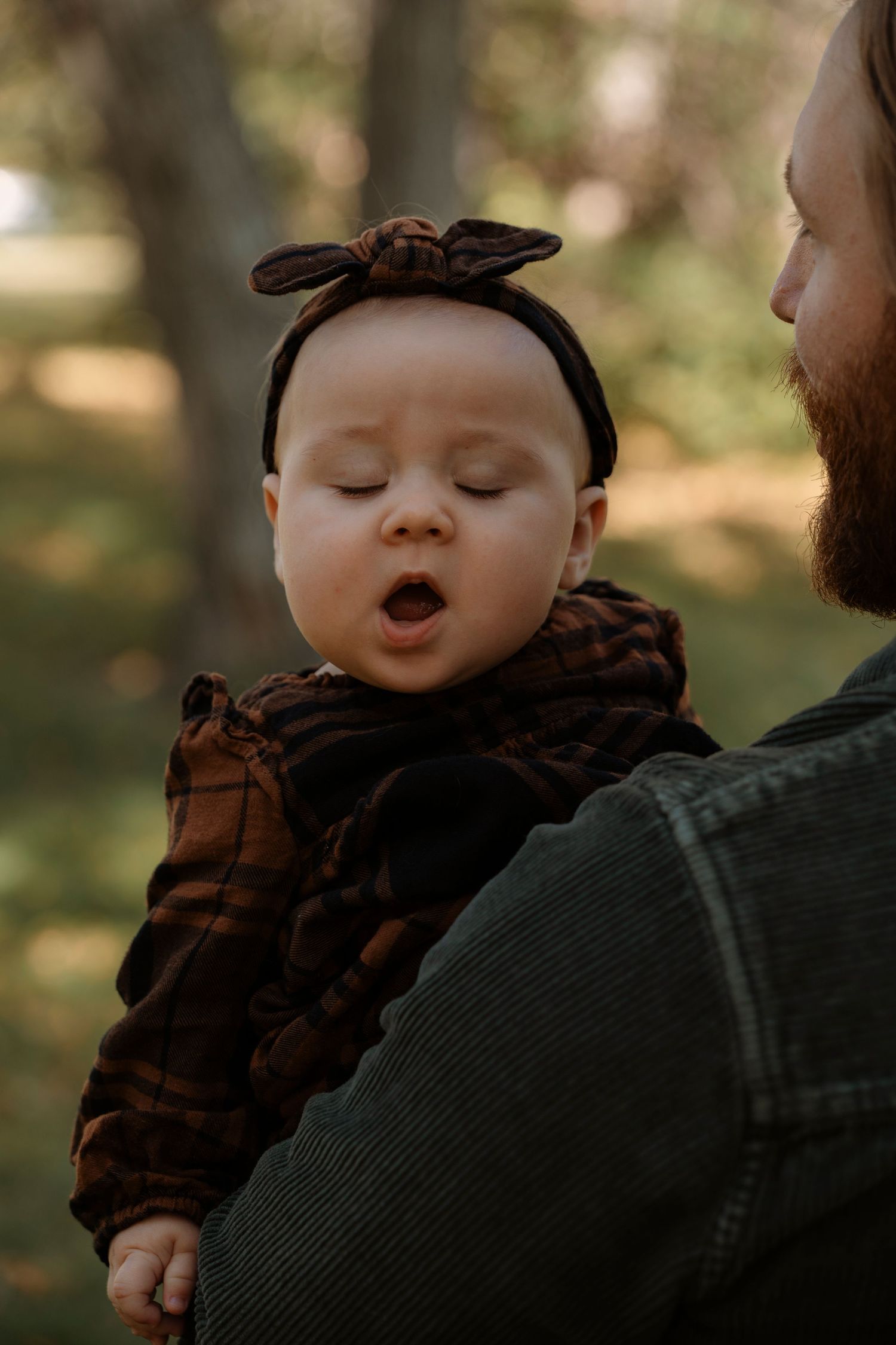A bearded man holds a sleepy baby wearing a dark plaid outfit and bow headband outdoors.