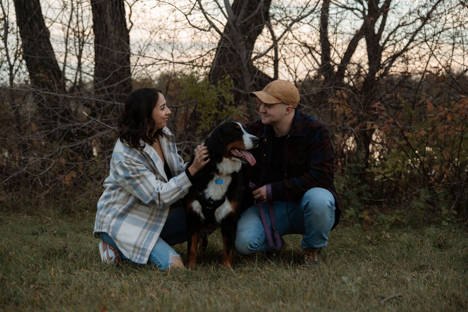 A couple crouching outdoors with their Bernese Mountain Dog amid autumn trees at dusk.