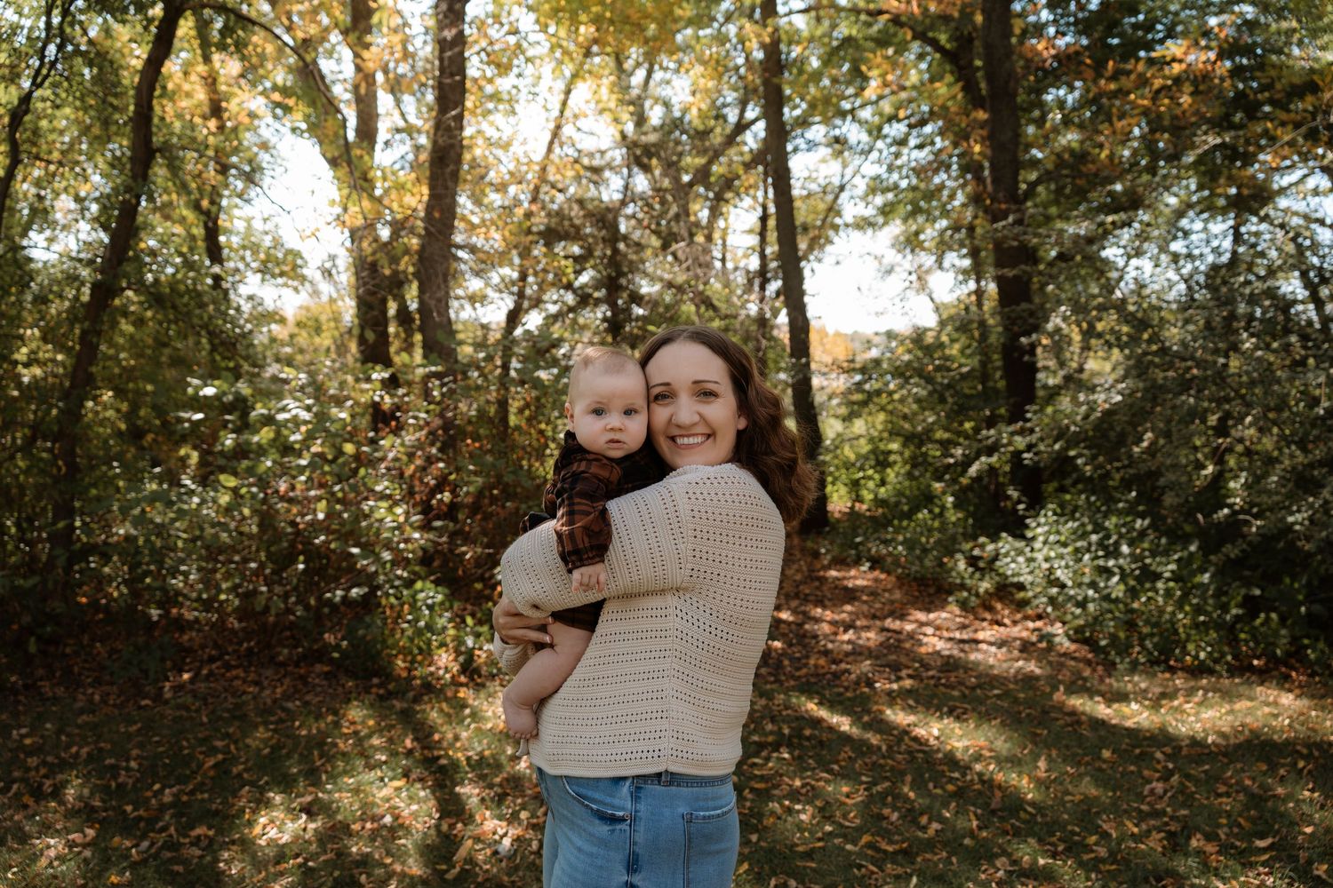 A smiling woman holds a baby outdoors in a sunlit autumn forest with golden and green foliage.