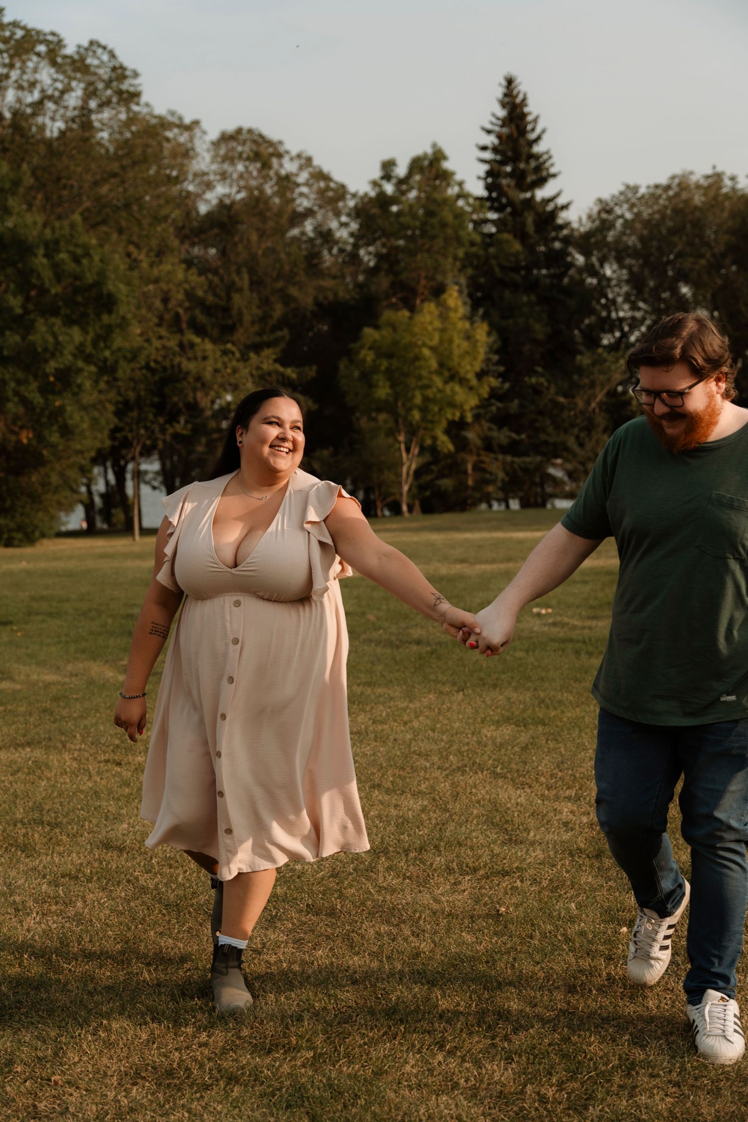 A happy couple holds hands while walking through a sunny green park, woman in a light pink dress smiling joyfully.