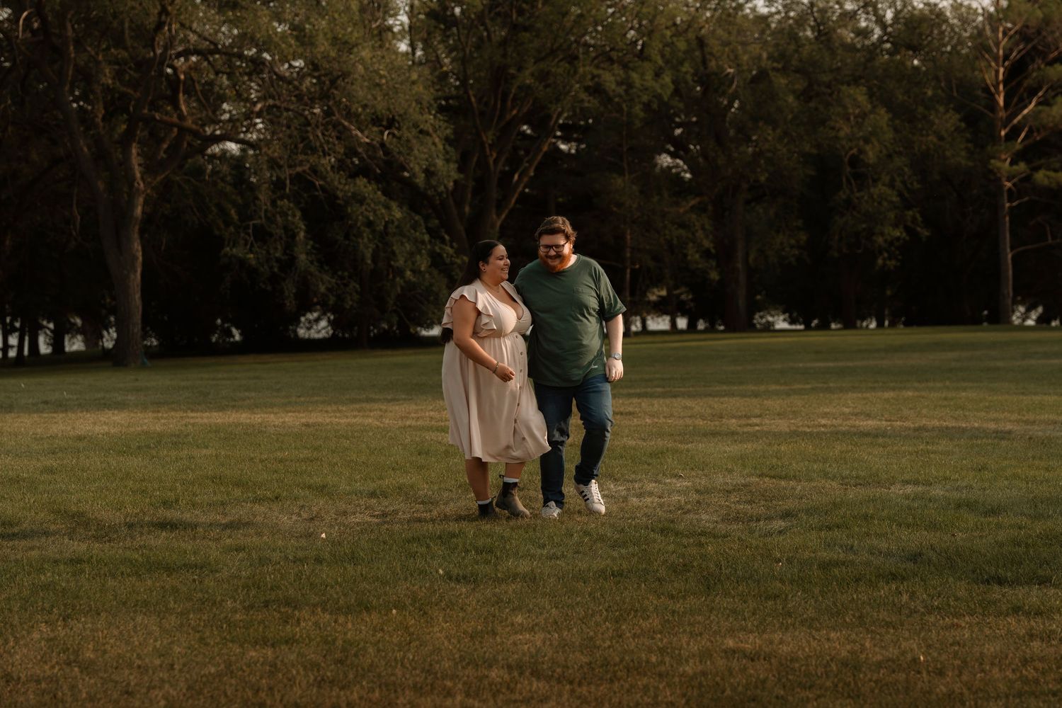 Pregnant couple walking hand in hand in a lush green park during golden hour.
