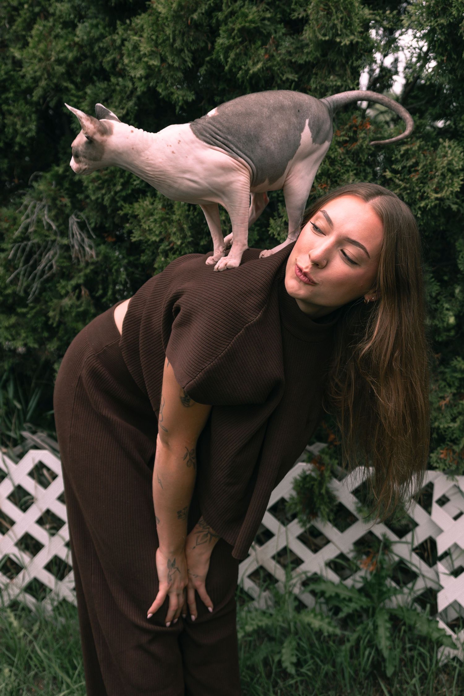 A young woman in a brown outfit balances a hairless Sphynx cat on her shoulders outdoors near green shrubs.