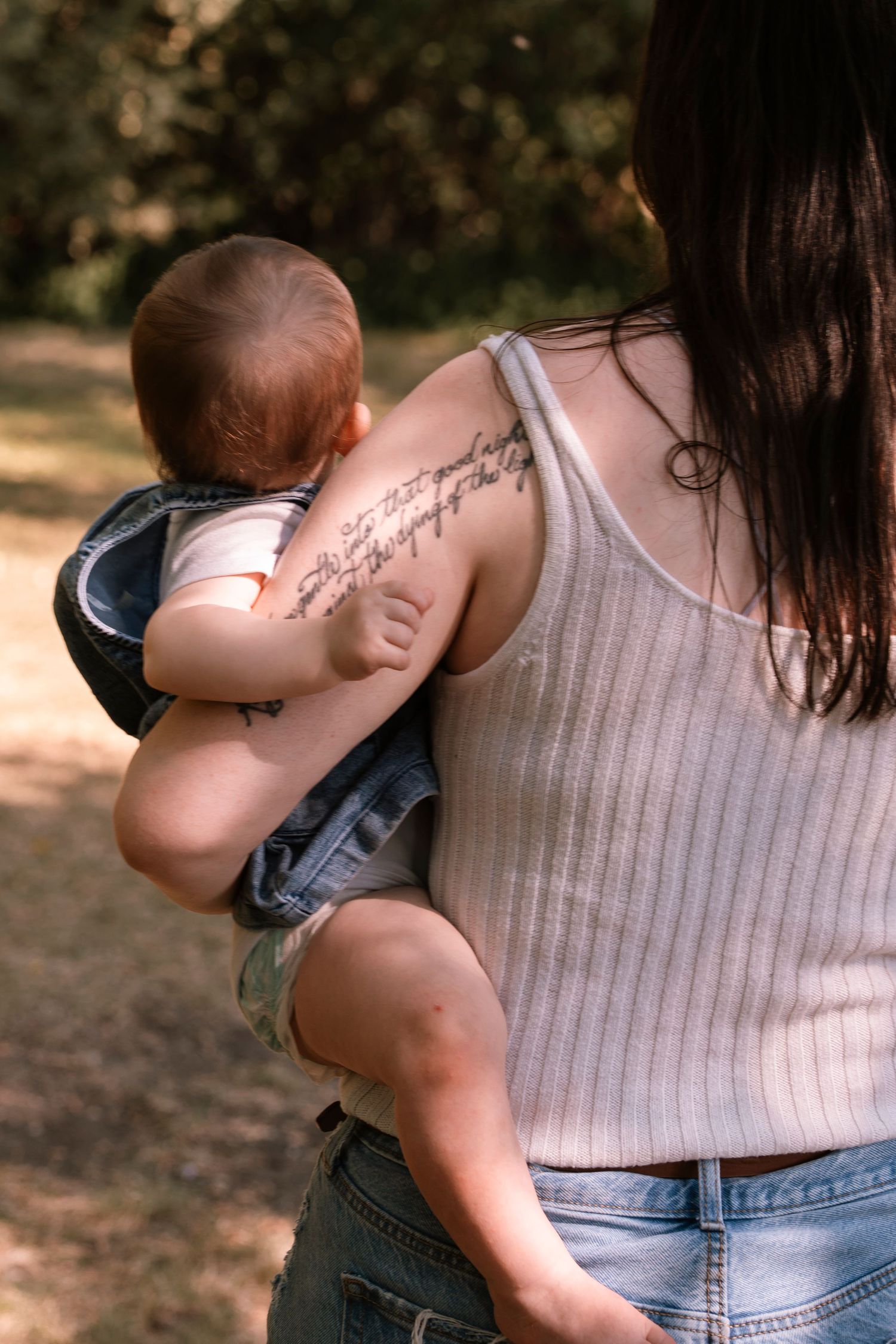 A parent in a white tank top holds a baby wearing denim overalls while standing outdoors in natural light. Regina Saskatchewan Family.