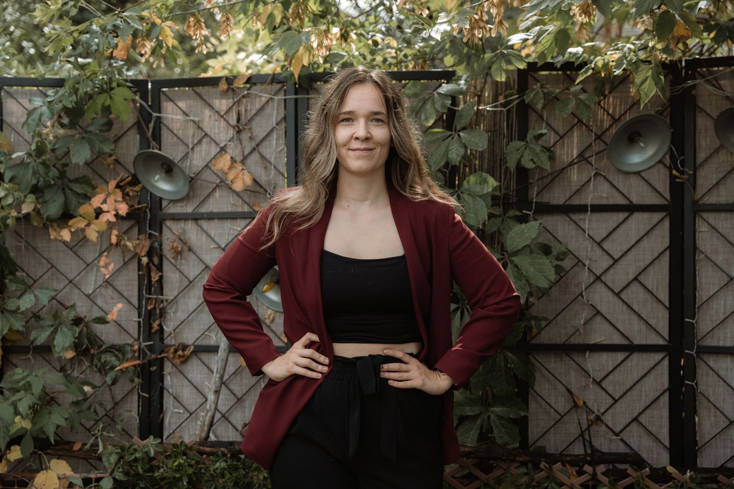 A person wearing a burgundy blazer and black dress poses outdoors against a wooden lattice fence with green foliage.