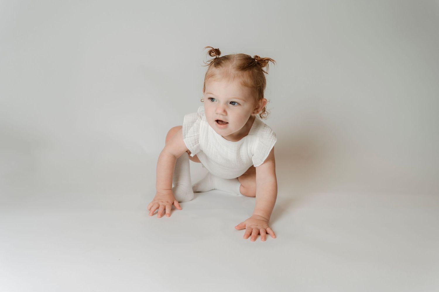 Adorable baby girl in white romper crawling on white floor with pigtails, studio portrait photography.