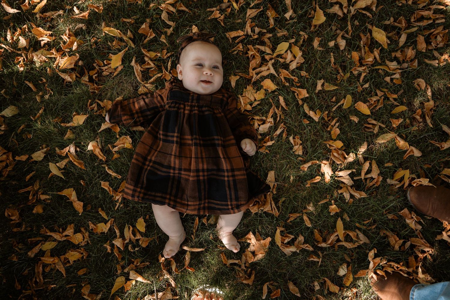 Baby in plaid dress lying on autumn leaves outdoors.