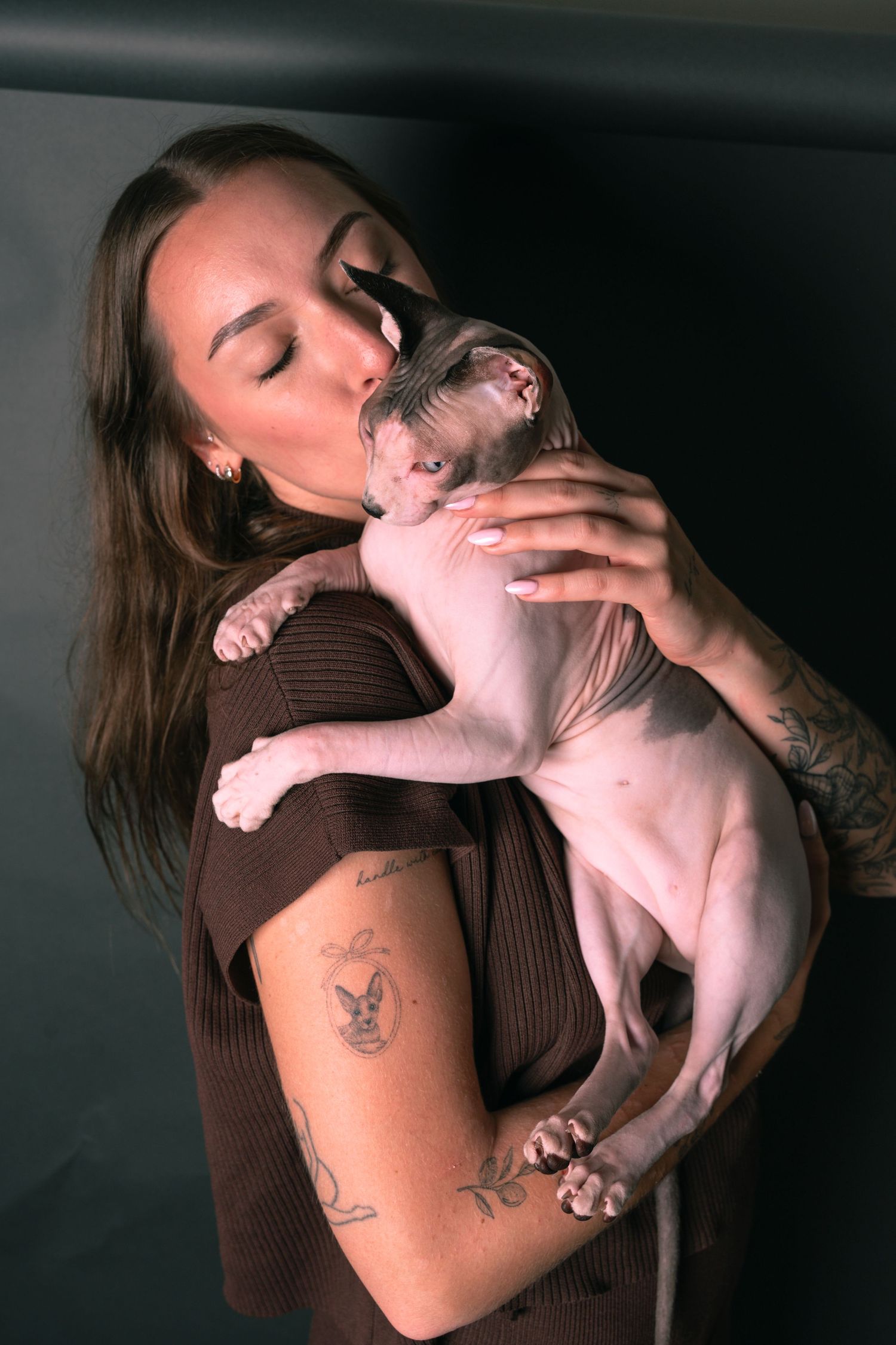 Woman with tattoos kissing a hairless Sphynx cat while holding it gently against her shoulder.
