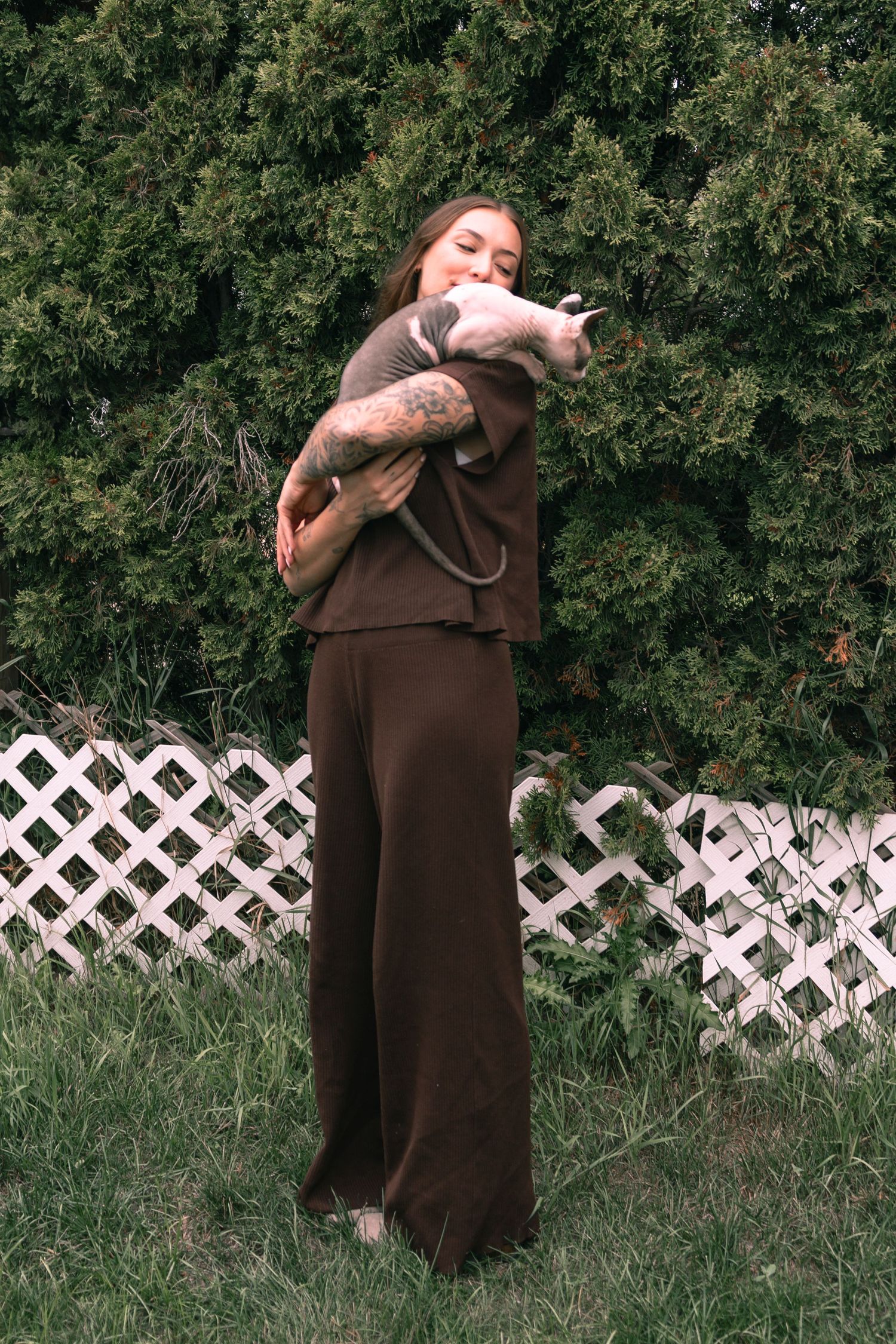 Woman in brown outfit holding a hairless Sphynx cat outdoors near green bushes and white lattice fence.