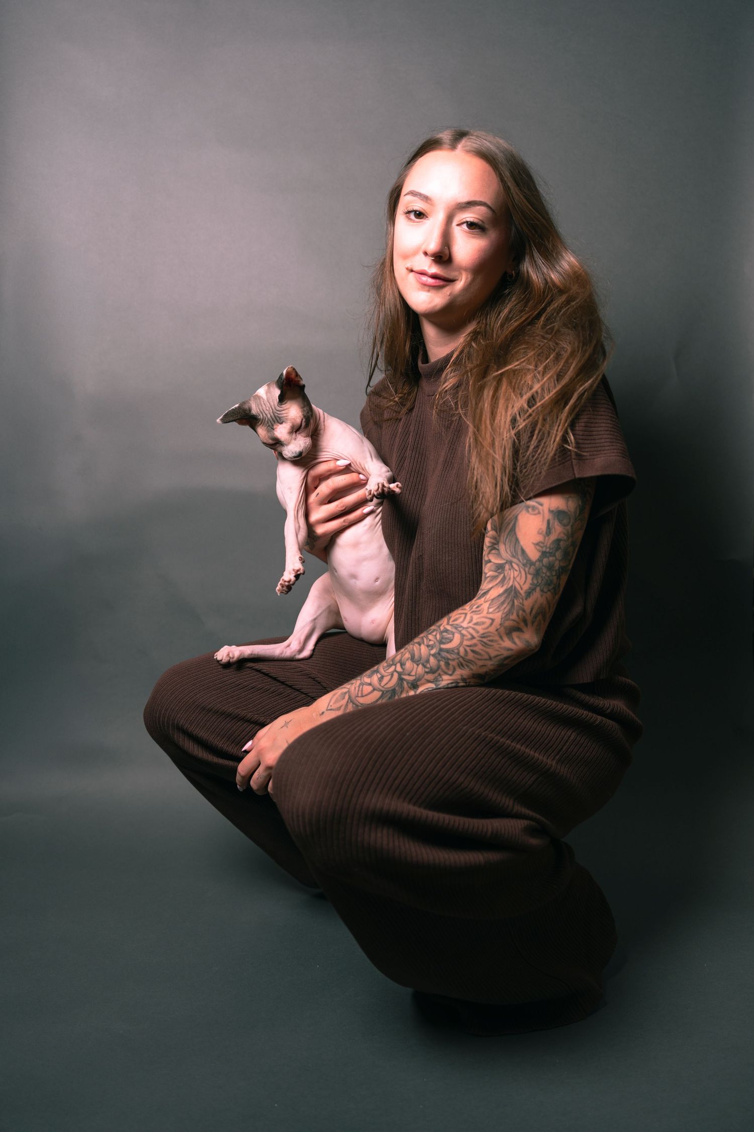 Woman with tattoos in brown outfit holds a hairless Sphynx cat while posing on gray studio background.