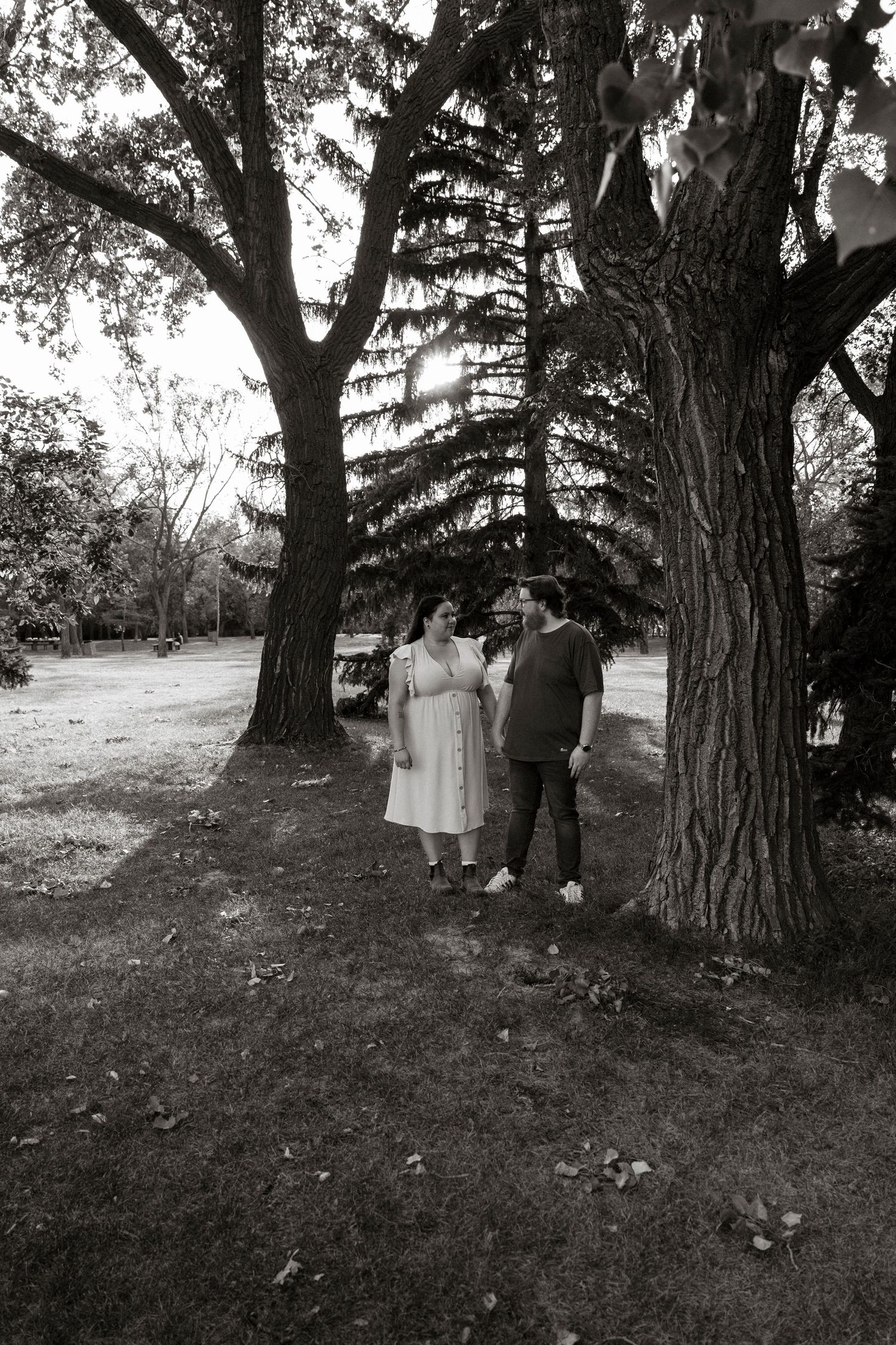 Black and white photo of a couple standing together under large trees in a park setting.