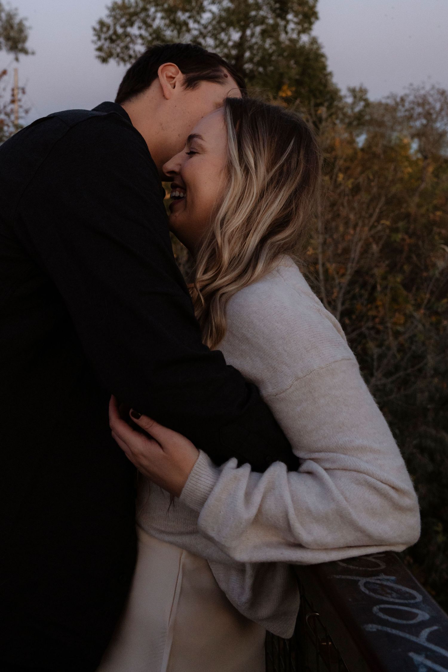 A couple shares a romantic embrace outdoors at dusk, the woman smiling in a cream sweater as they lean in for a kiss.